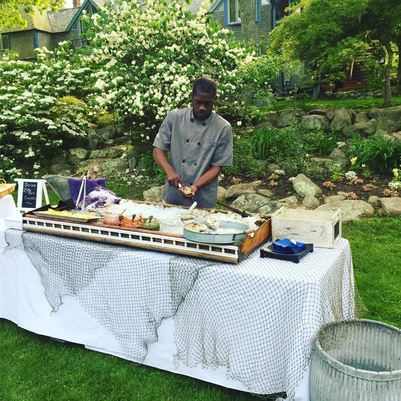 rawbar shucker preparing seafood on an outdoor buffet table with garden and blooming trees in the background.