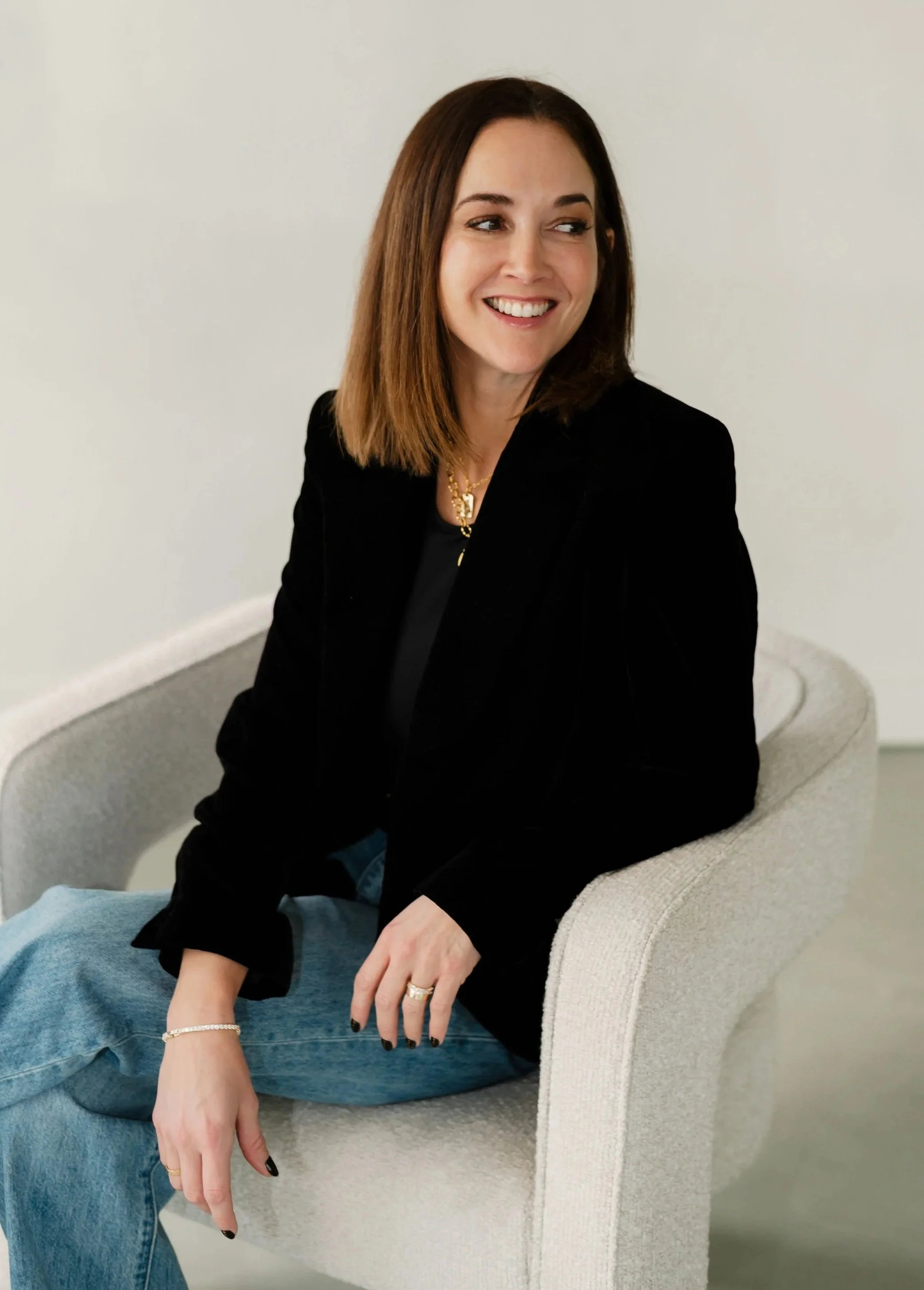 A woman with shoulder-length brown hair smiling, seated on a beige curved chair against a plain white wall, wearing a black blazer, gold jewelry, and casual blue jeans.