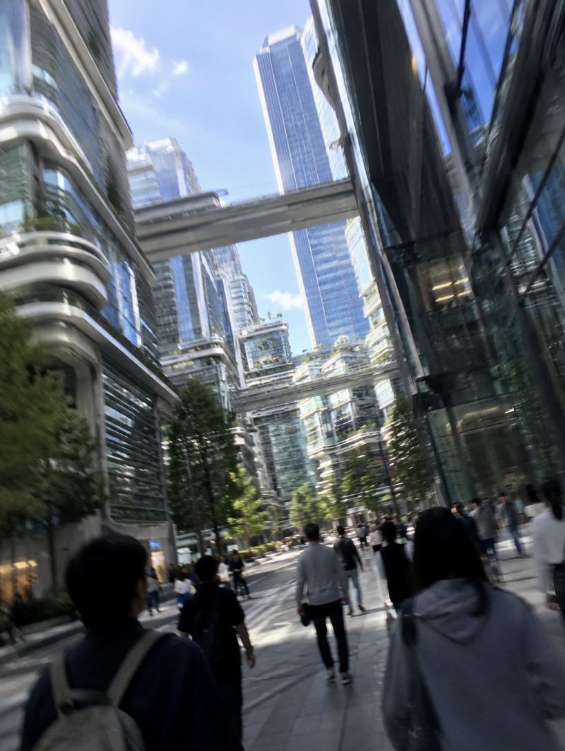 City street scene with modern glass buildings, people walking, and sky with clouds in an urban area.