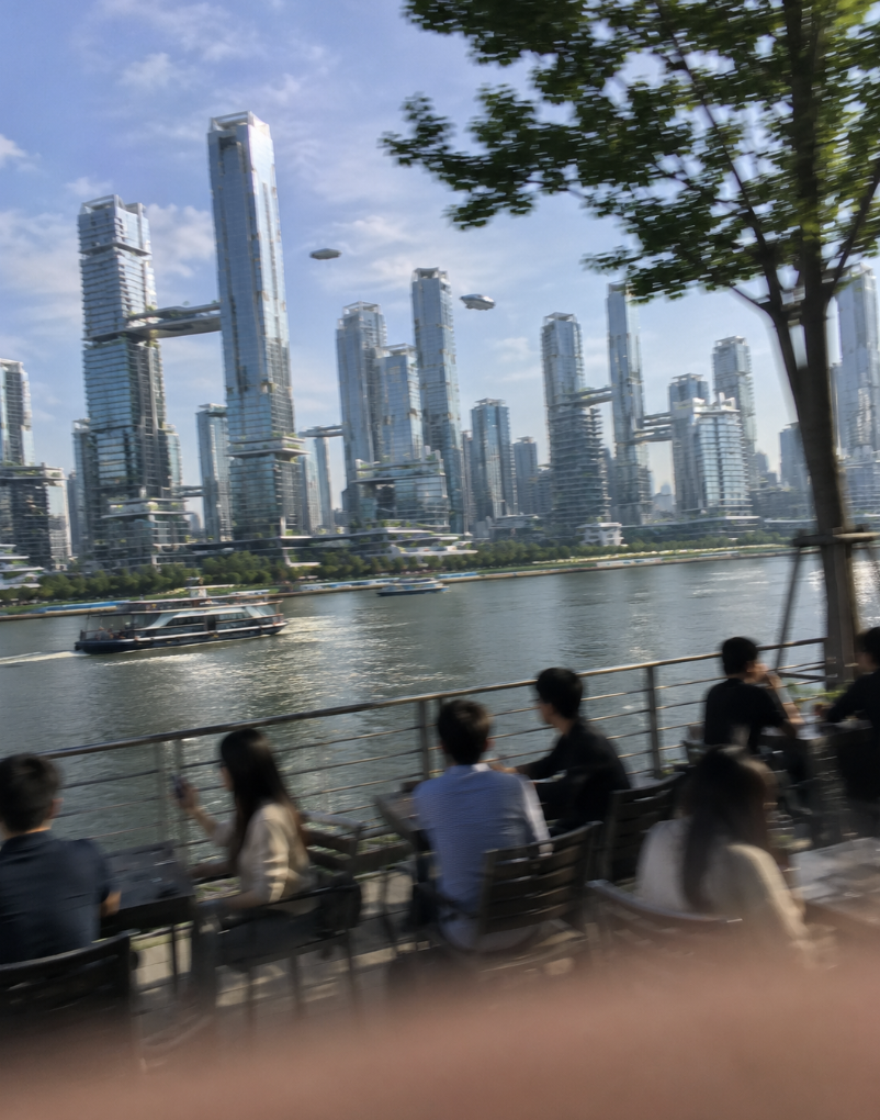 People sitting at outdoor tables by the river in a city with modern high-rise buildings and flying vehicles in the sky.