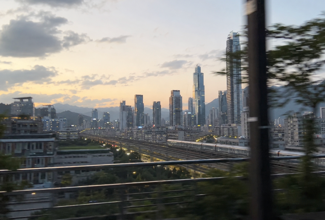 A city skyline at dusk with tall skyscrapers, a mountain range in the background, and blurred foreground elements including trees and railway tracks.