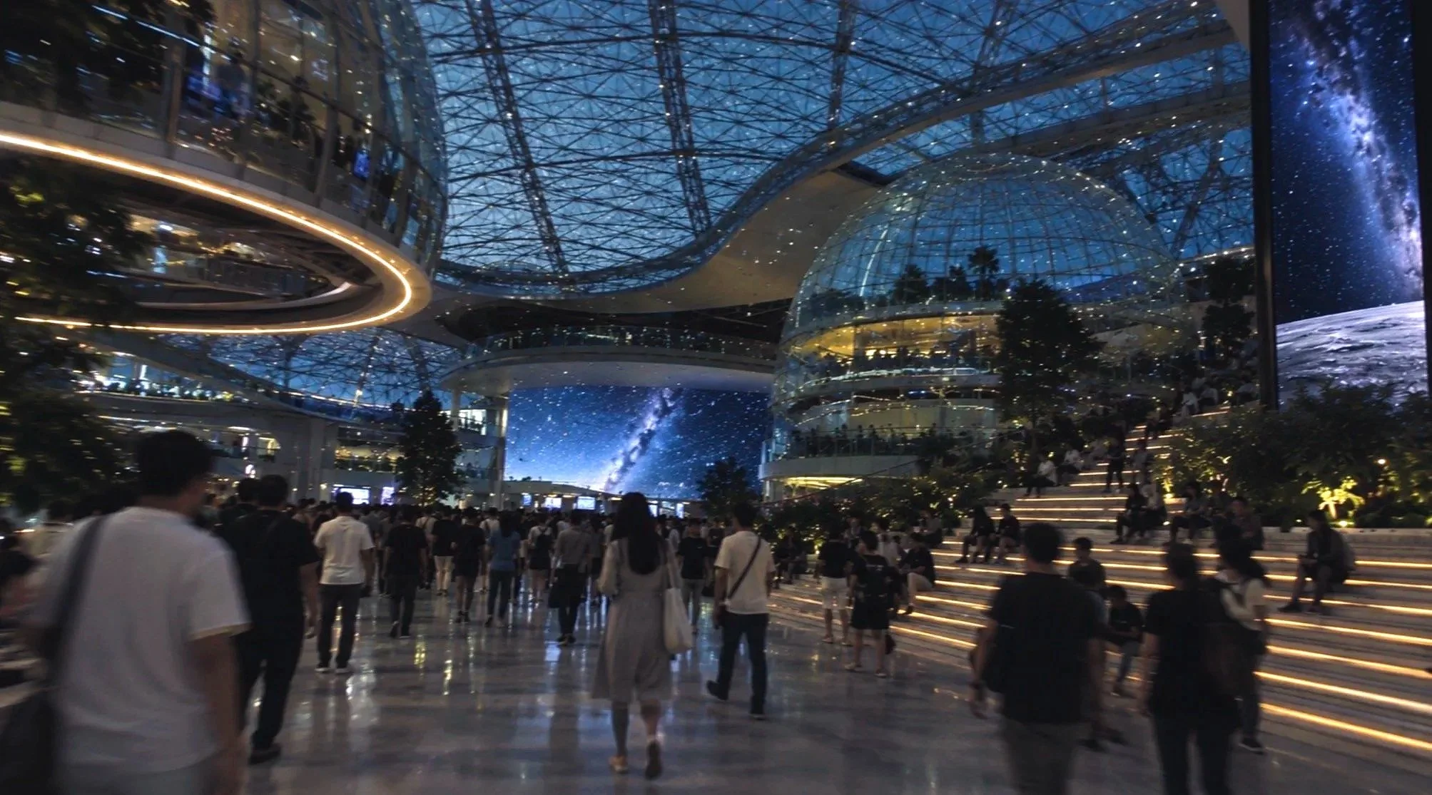 A busy indoor space with a glass ceiling and modern architecture, filled with people walking and sitting on illuminated steps. There are trees and a large digital screen, with a starry night sky and galaxy images projected on the walls.