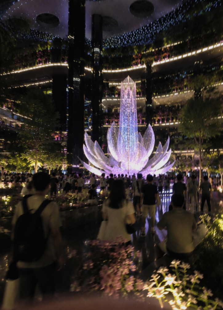 Indoor garden with a large illuminated lotus flower sculpture at the center, surrounded by people, with reflective floors, greenery, and hanging blue fairy lights.