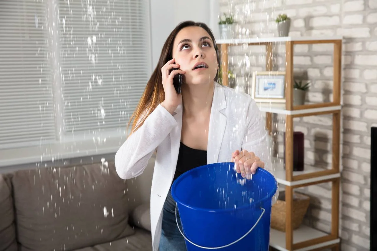 A woman, inside a living room with water falling from the ceiling, is holding a blue bucket catching some of the water. She is on the phone.