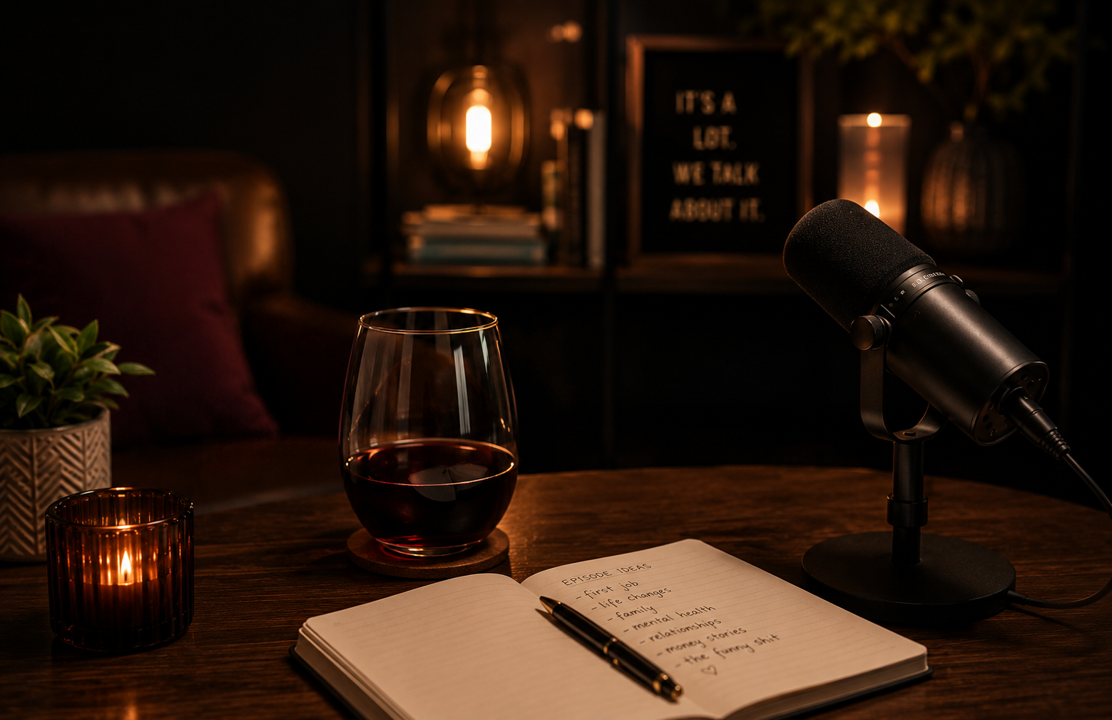 A cozy, dimly lit recording setup featuring a laptop, a microphone on a stand, a glass of red wine, a lit candle, a potted plant, and a notebook with handwritten notes. In the background, there is a bookshelf with books, candles, and a sign that reads 'It's a lot. We talk about it.'
