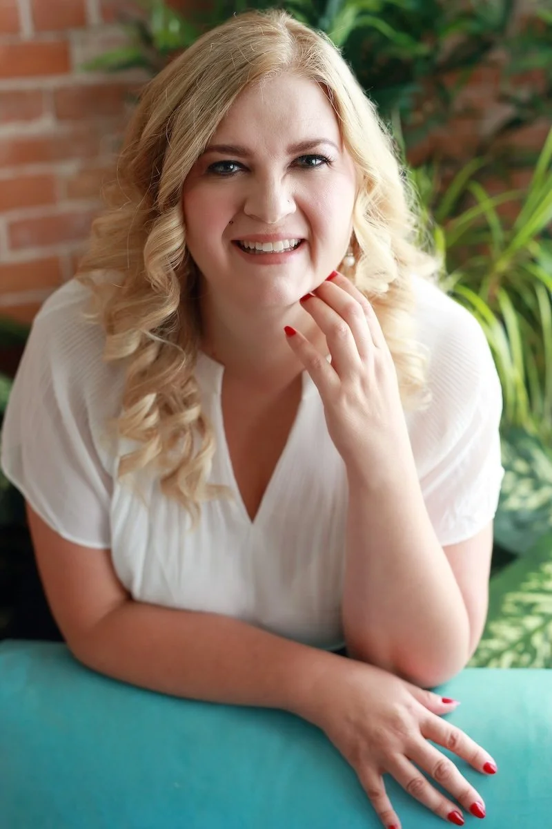 A smiling blonde woman with curly hair, wearing a white top, sitting at a table with a teal surface, touching her chin and looking at the camera. In the background, there are green plants and a brick wall.