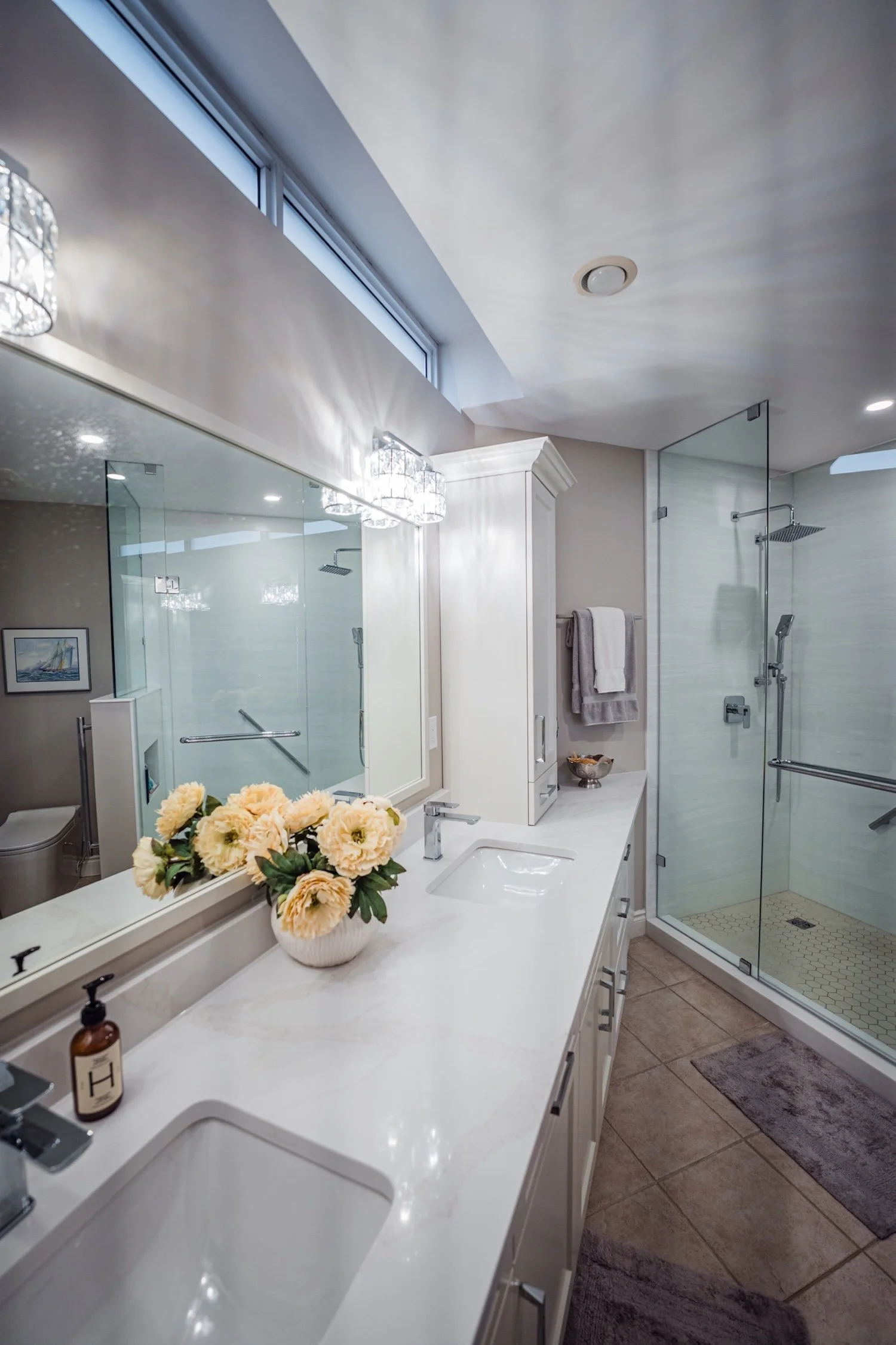 A bathroom with a large mirror above a white countertop, a sink, a vase with yellow flowers, and a shower with glass doors, a towel hanging on a hook, and a small shower bench.