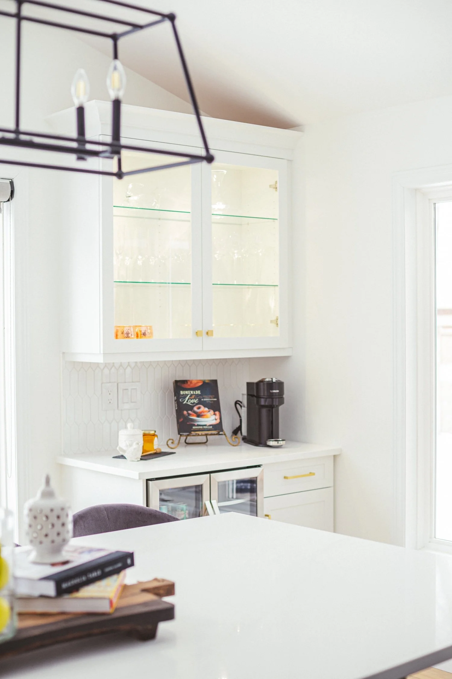 A white kitchen with a white cabinet, black coffee maker, books, and a purple cushion on a chair.