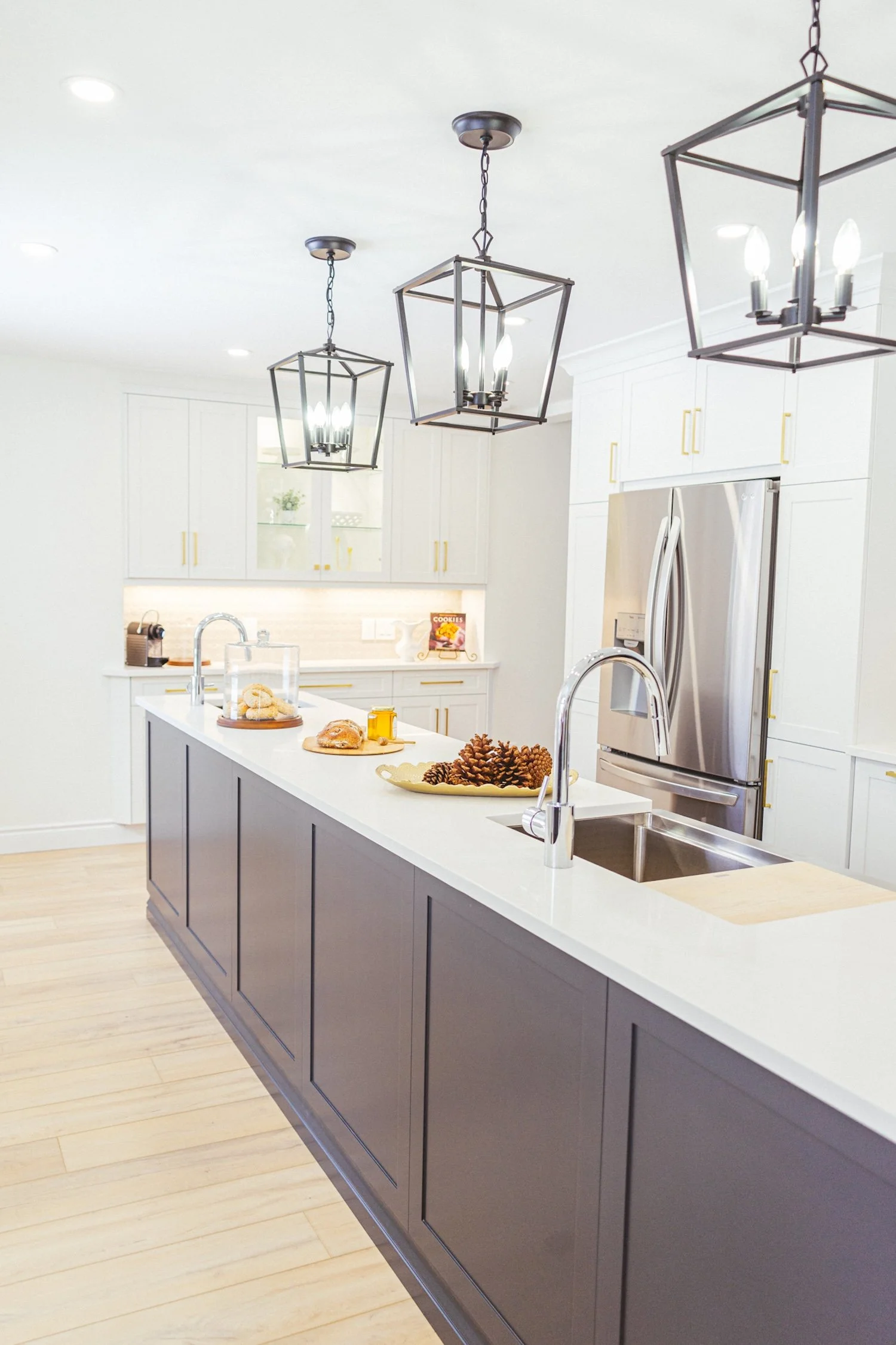 Modern kitchen with white upper cabinets, a gray island, and three geometric pendant light fixtures hanging from the ceiling. The kitchen features a stainless steel refrigerator and a white countertop with baked goods and a tray of pinecones.