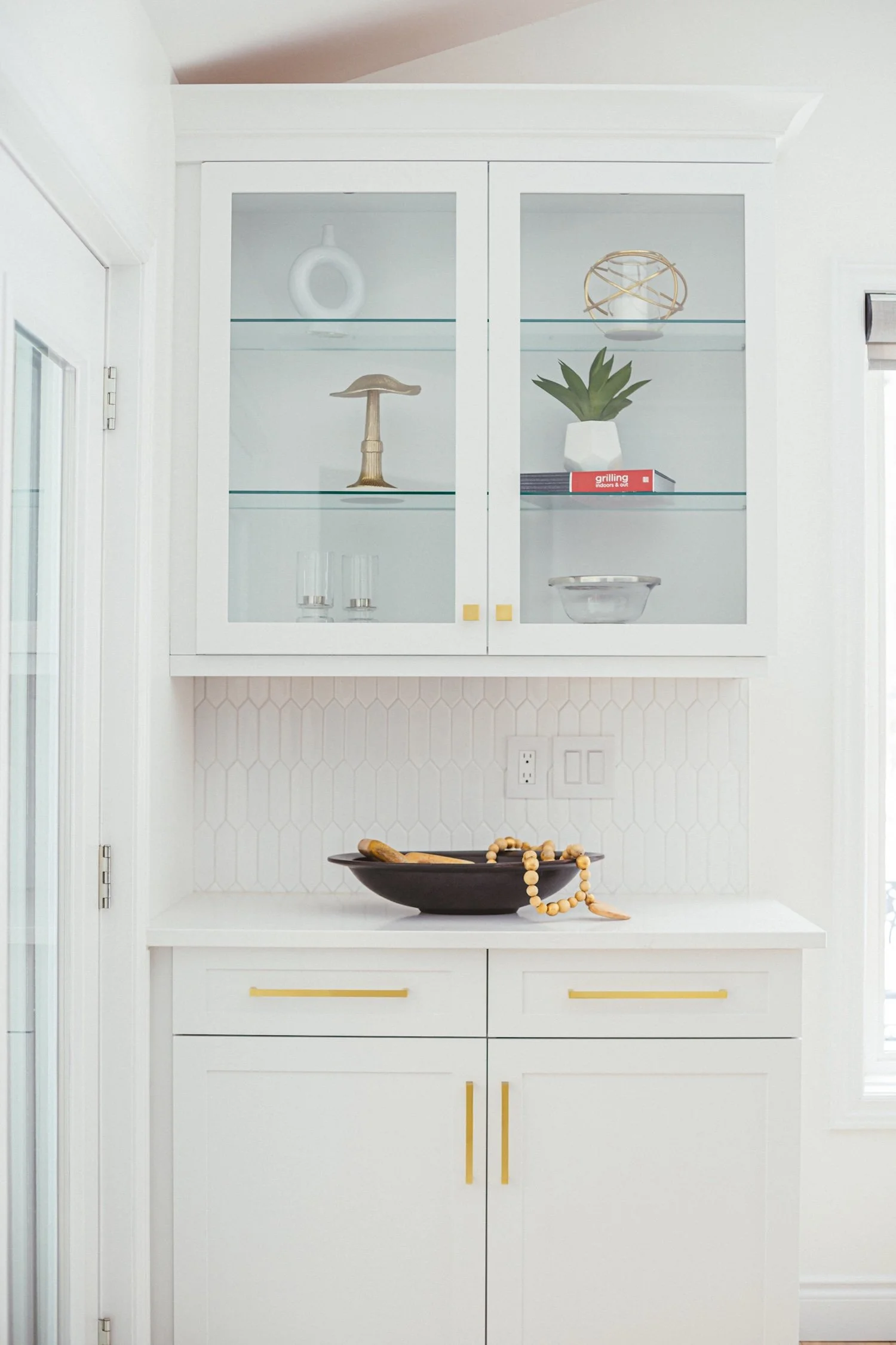 A white kitchen cabinet with glass doors displaying decorative items, a black tray with a wooden spoon and a beaded necklace, and white walls with a hexagonal tile pattern.