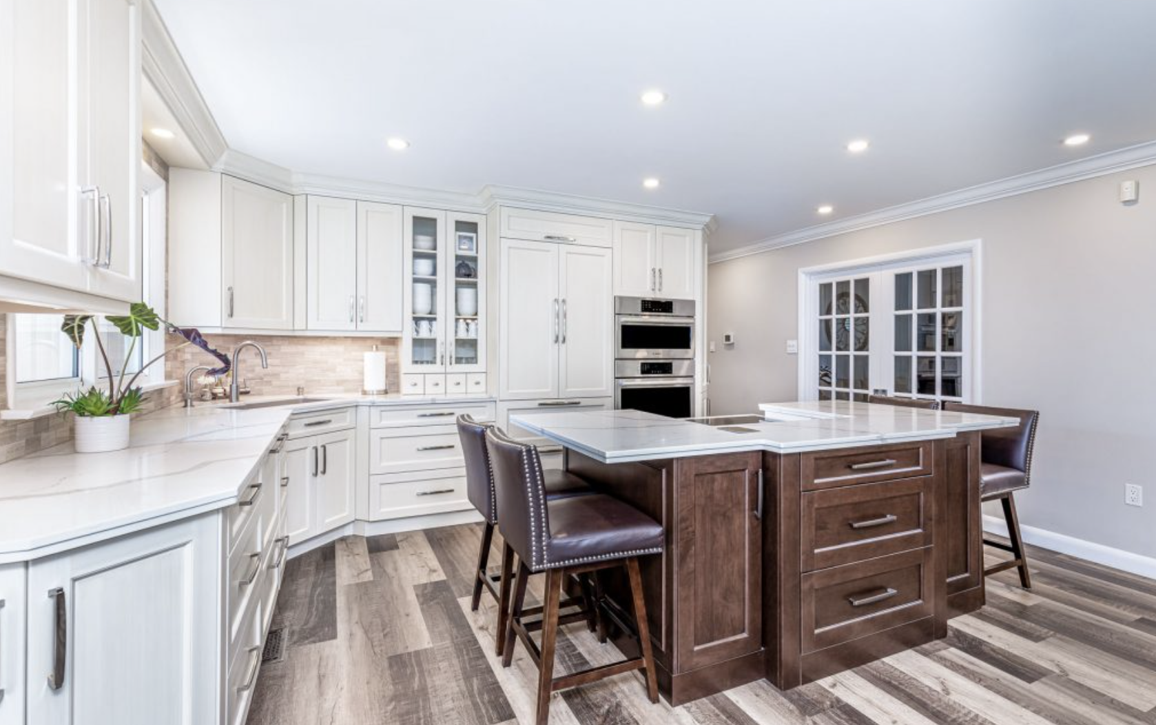 Modern kitchen with white cabinets, a wooden island, granite countertops, and stainless steel appliances. There are two barstools at the island, a potted plant near the window, and hardwood flooring.
