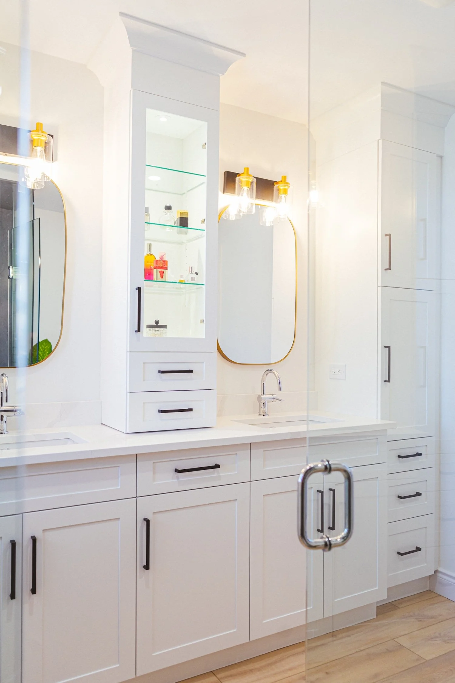 Modern white kitchen with a white countertop, cabinets with black handles, and a sink. There's a white tall cabinet with glass shelves displaying bottles, surrounded by wall mirrors with gold trim. A clothes-locked glass door reflects part of the room.