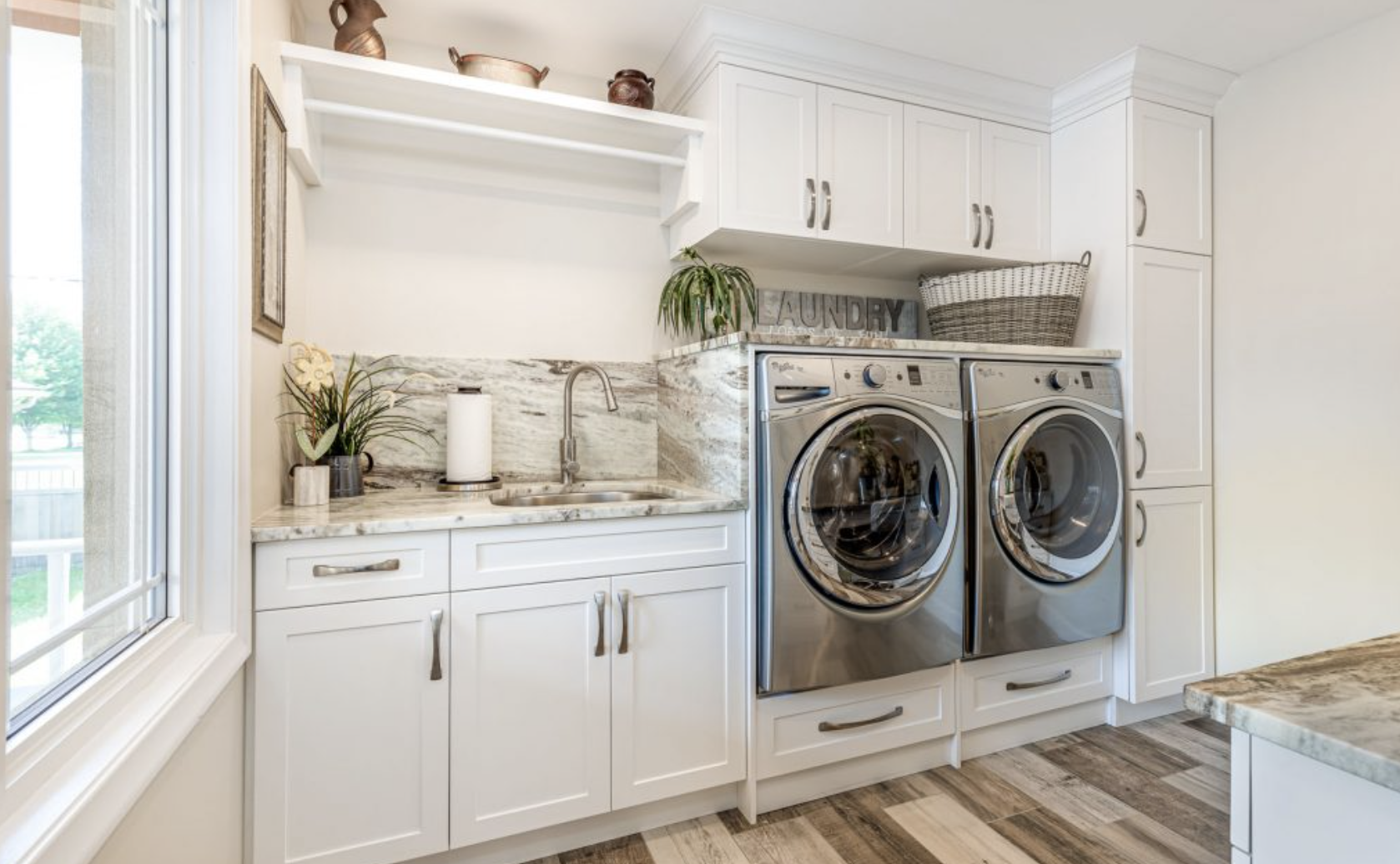 Laundry room with white cabinets, marble countertop, and a front-loading washer and dryer, with a wicker basket and a small plant on top.