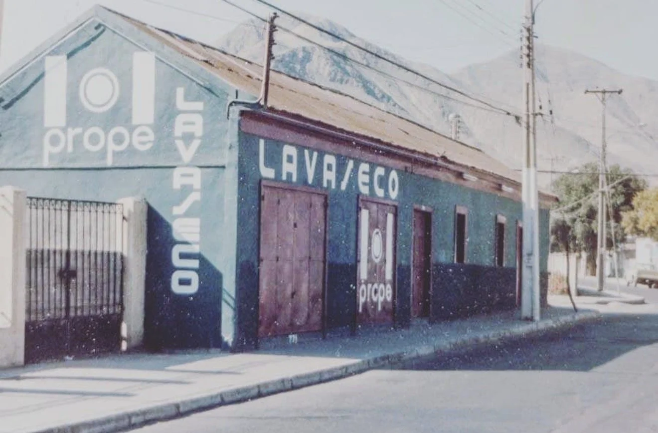 Edificio de ladrillos con señalización de lavandería y ropa en la fachada, en una esquina de la calle, con postes de electricidad y árboles en el fondo.