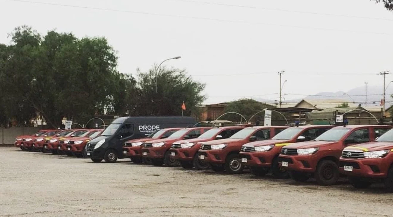 Lote de vehículos, principalmente camionetas rojas con algunos usados en color negro y otros en color blanco, en un estacionamiento al aire libre con árboles y estructuras al fondo.