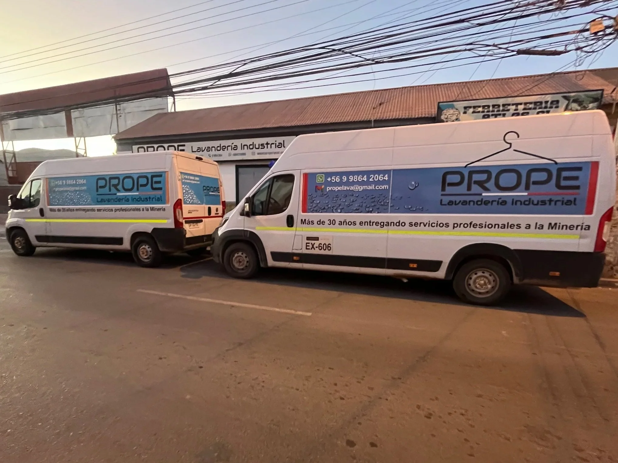 Dos furgones blancos con publicidad de una lavandería industrial en color azul, rojo y negro estacionados en la calle frente a un edificio que también tiene cartel de lavandería, en una vía urbana, durante el amanecer.