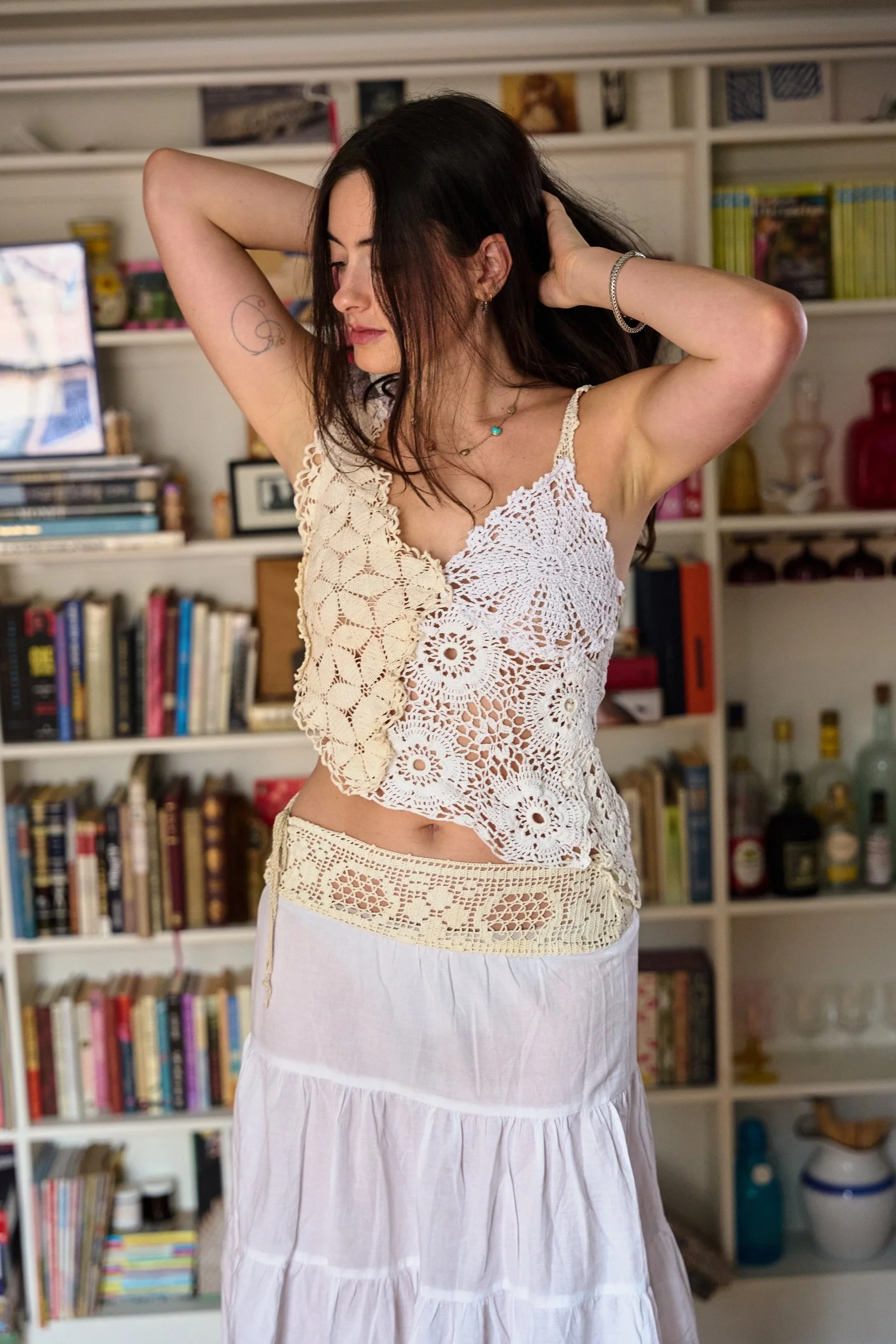 A woman with long dark hair, wearing a cream-colored crocheted top and a white skirt, standing in front of a bookshelf with books and decorative items, with her arms raised behind her head.