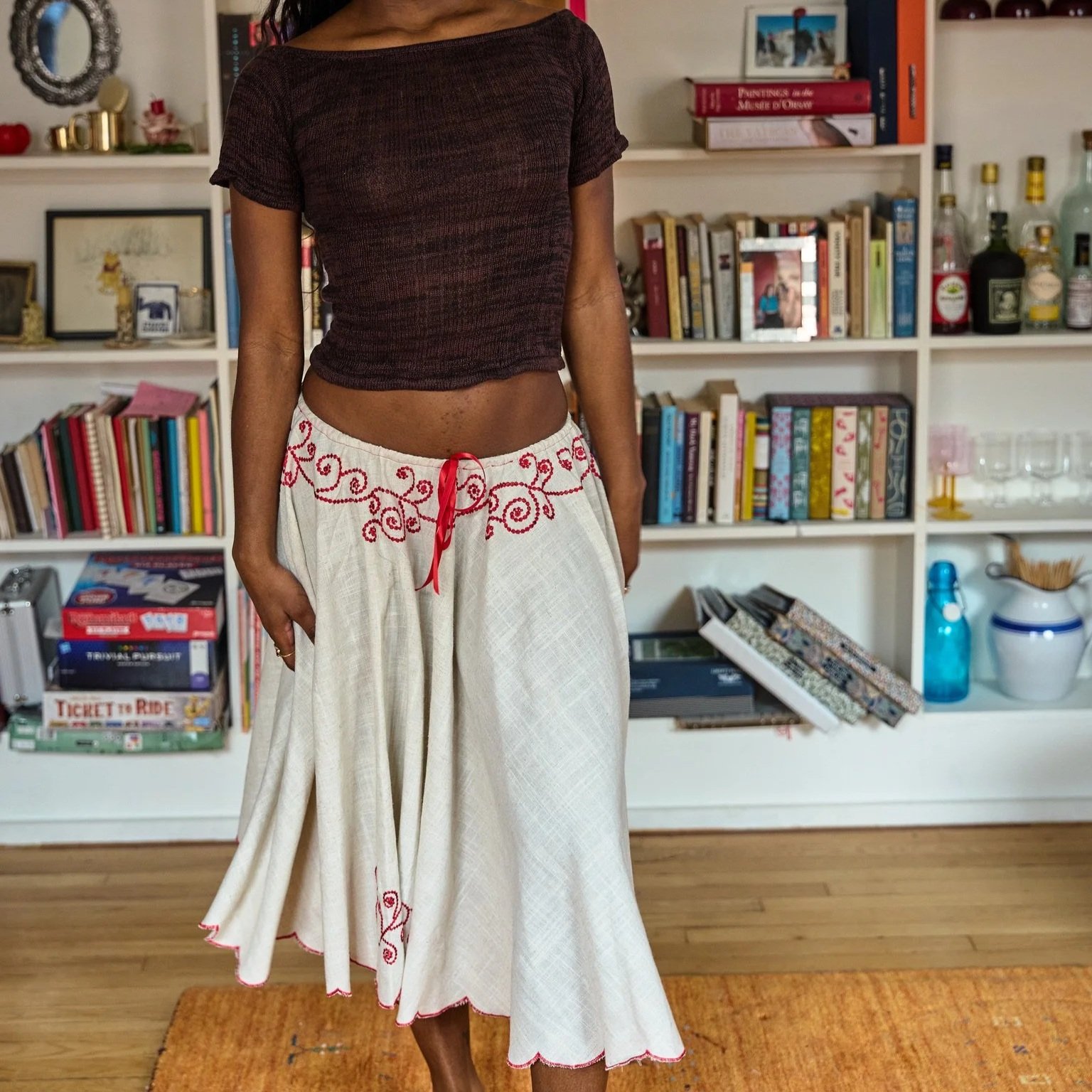 A woman wearing a dark brown top and a long white skirt with red embroidery, standing in a room with a white bookshelf filled with books, picture frames, and decorative items.