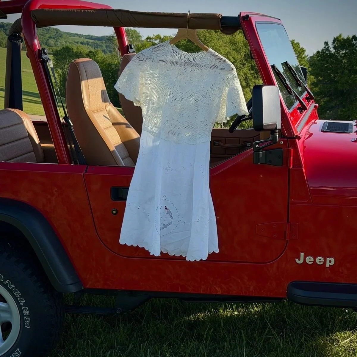 A white dress hangs on a hanger attached to the inside of a red Jeep, with the background of a rural landscape with green trees and grass.