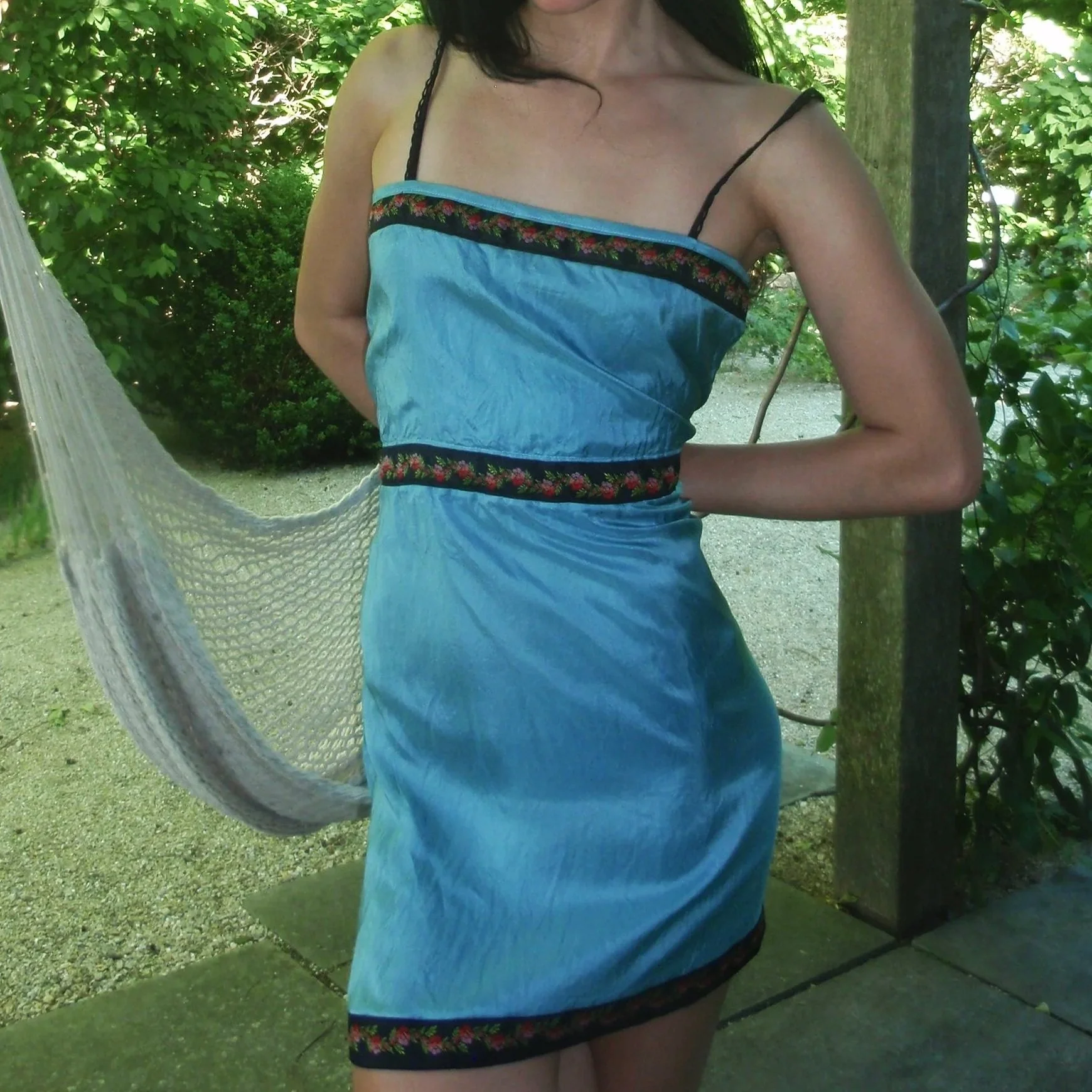 A woman in a blue dress with black and floral embroidered trim standing outdoors near a tree and a hammock, with lush green foliage and sunlight in the background.