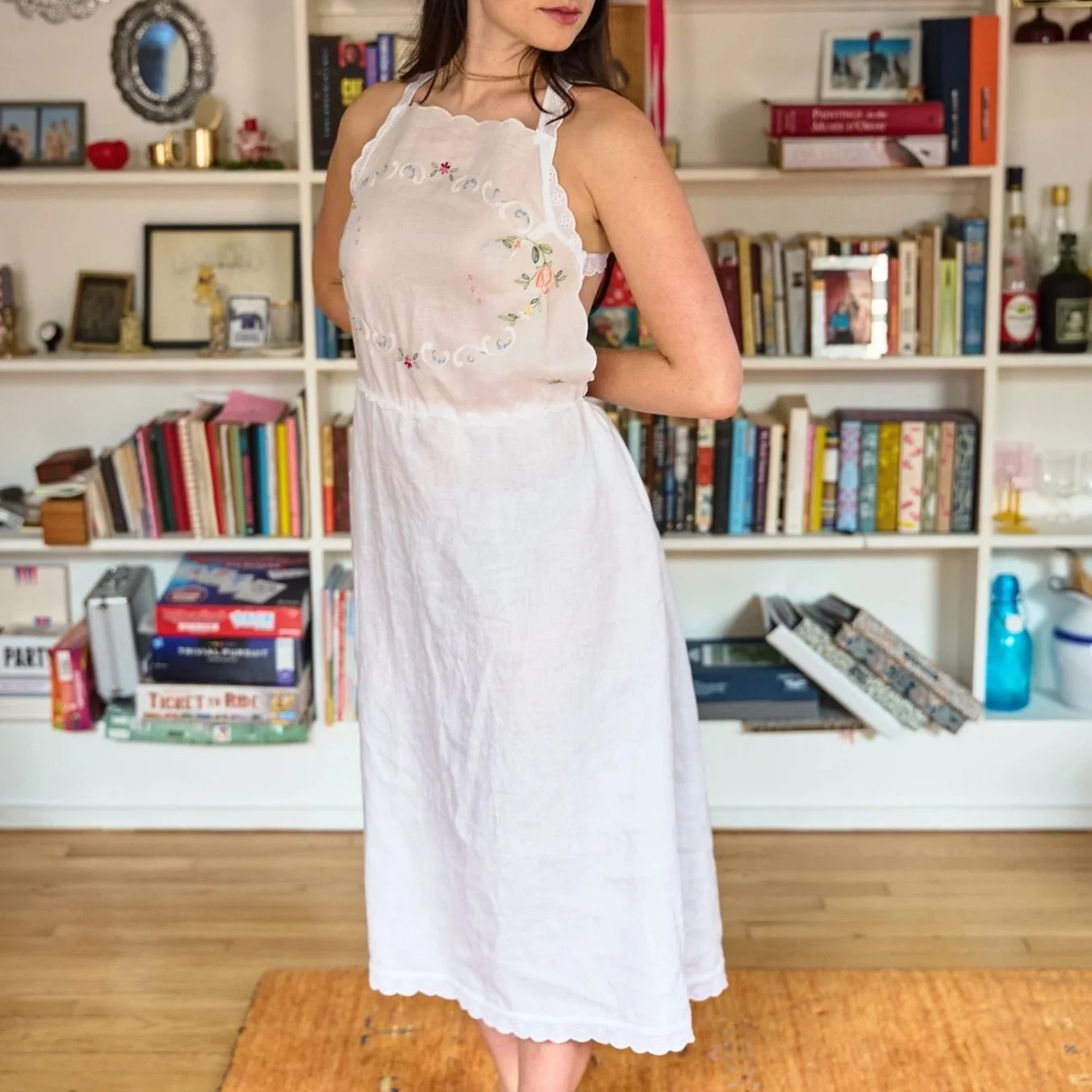 A woman wearing a white embroidery apron standing in a room with a bookshelf filled with books and decorative items. The room has wooden flooring and a small orange rug.