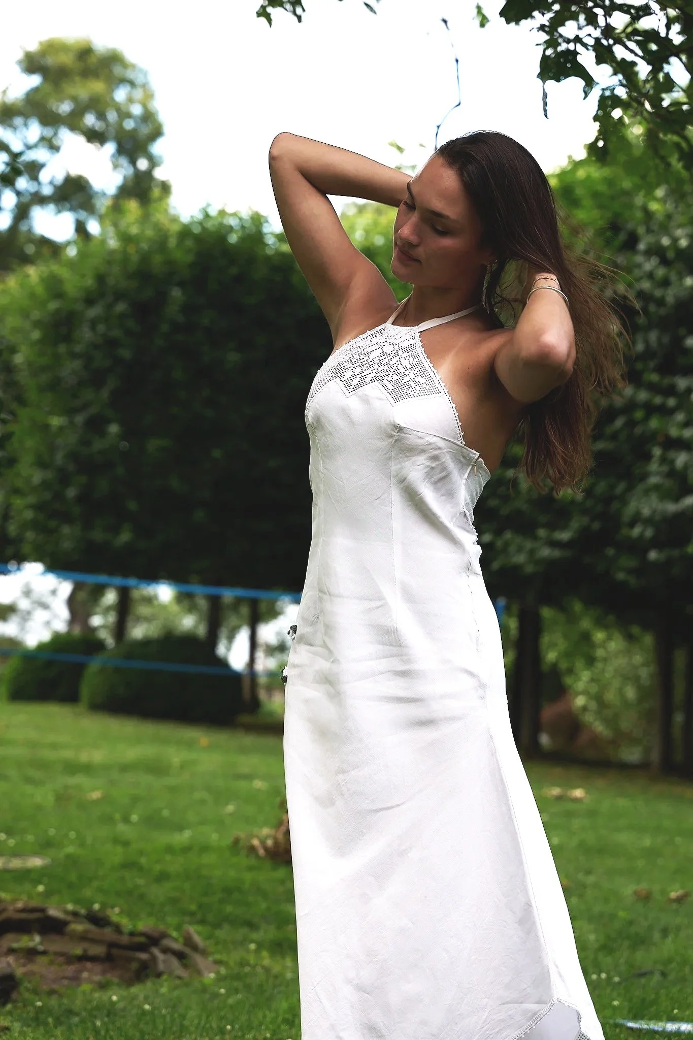 Woman in a white dress standing outdoors in a grassy park with green trees in the background, her eyes closed and arms raised behind her head.