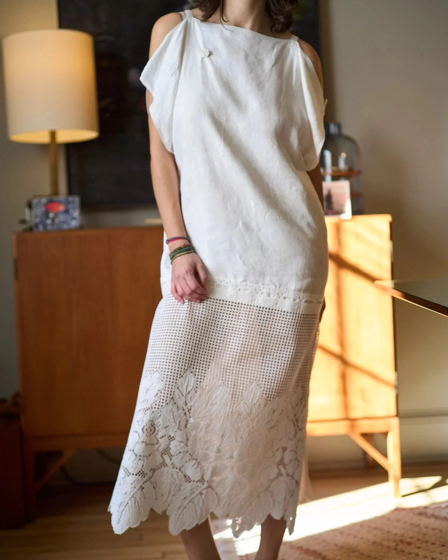 Woman wearing a white, lace dress standing in a room with warm lighting, next to a dresser and a lamp.