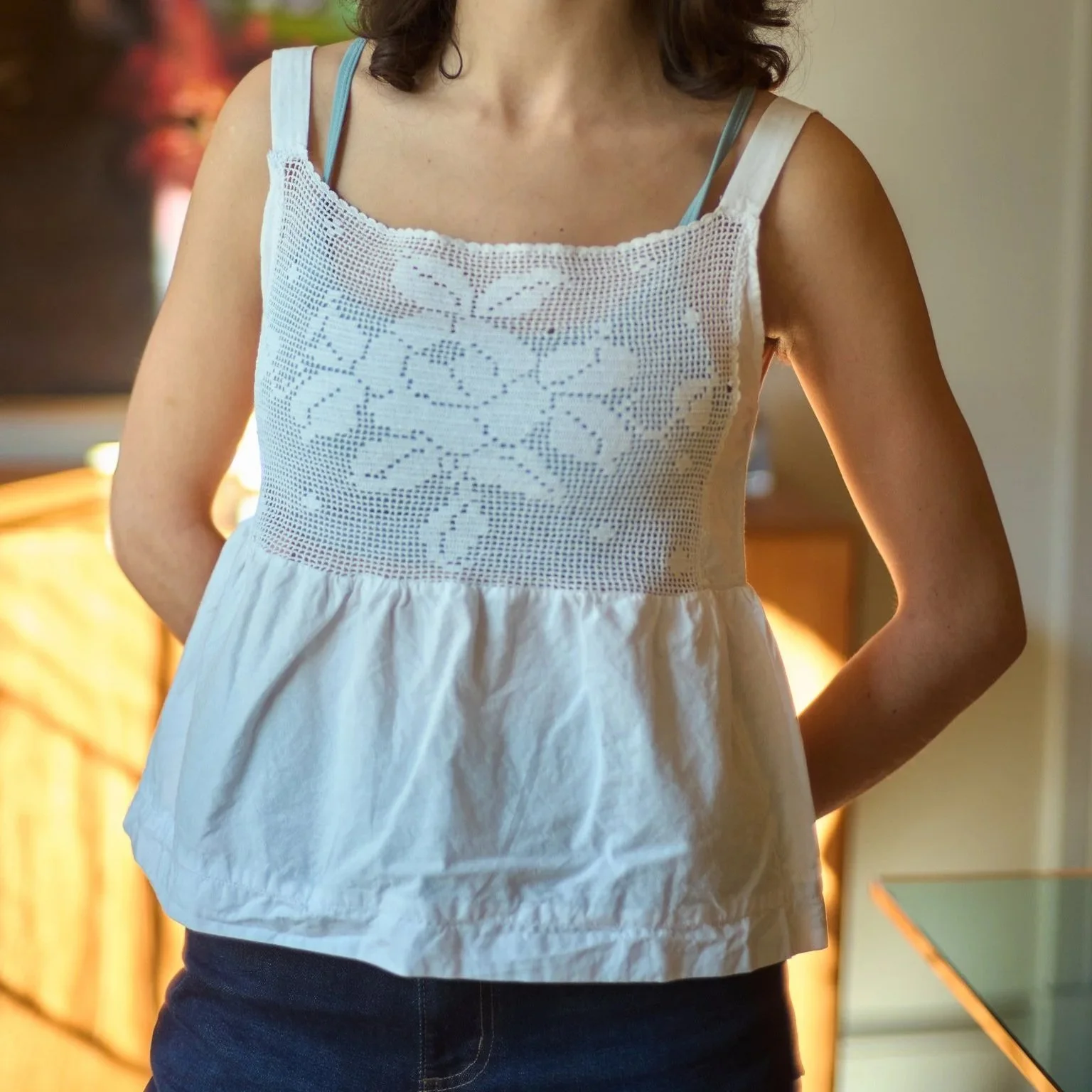 A woman wearing a white sleeveless dress with a lace floral pattern on the front, standing indoors.