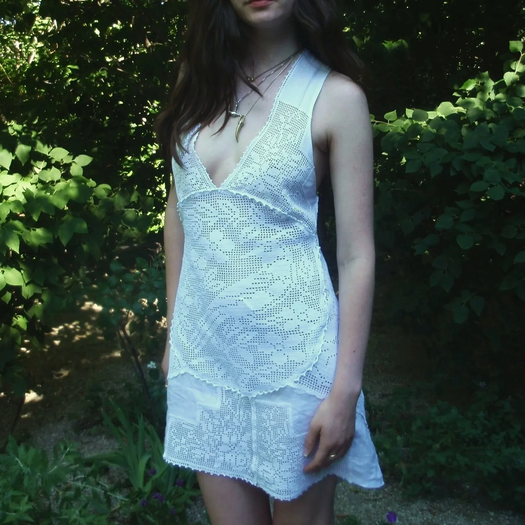 A woman wearing a white, lace-patterned dress with layered necklaces in front of green foliage.