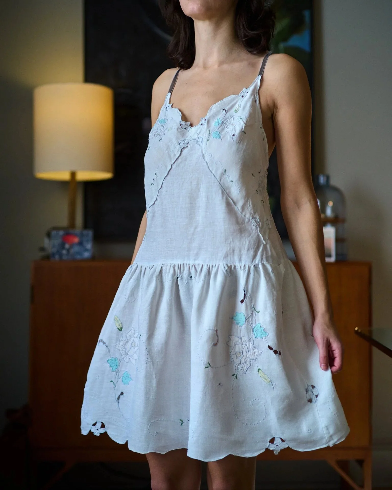 A woman wearing a sleeveless, white, embroidered dress with floral designs, standing indoors in front of a wooden cabinet, with a lamp and picture frame in the background.