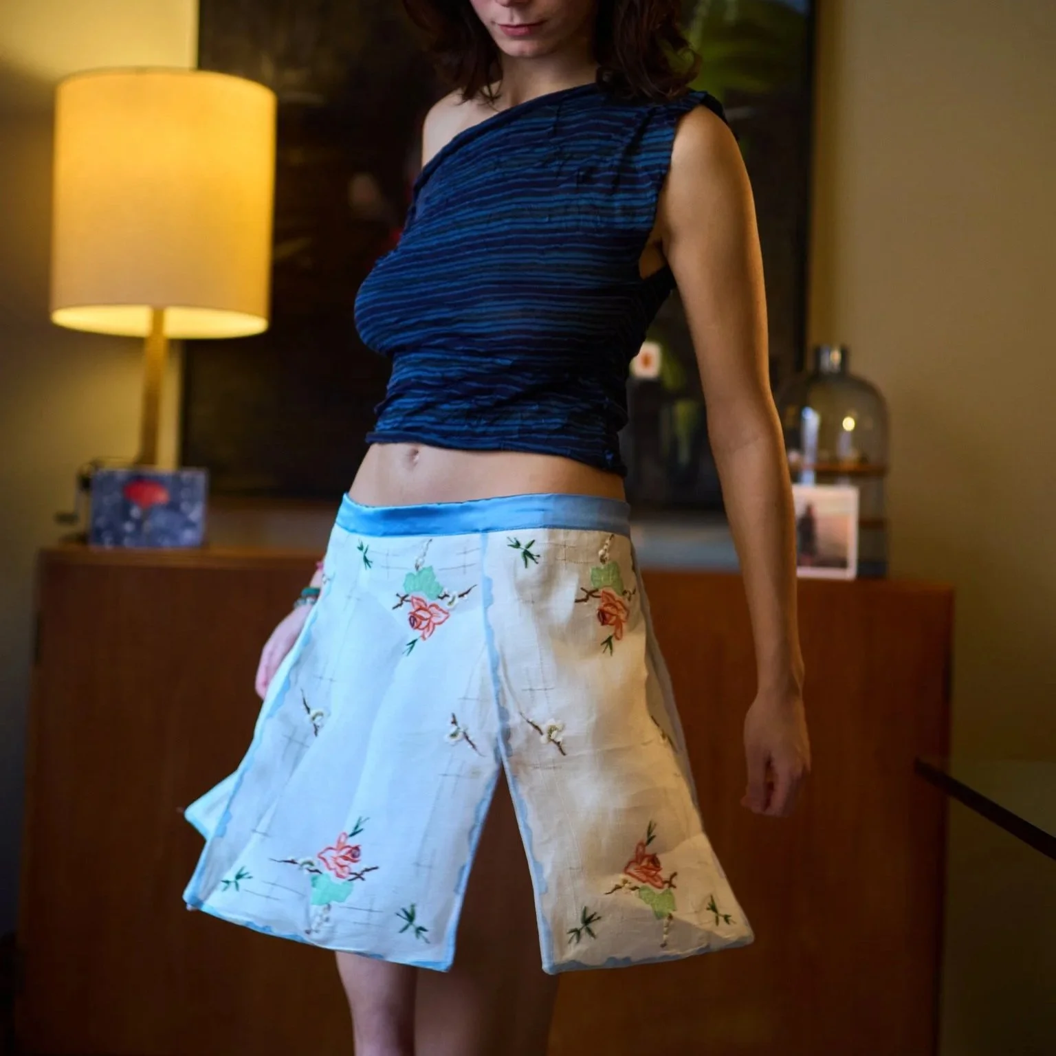 A woman standing indoors wearing a blue sleeveless top and white pants with a floral pattern.