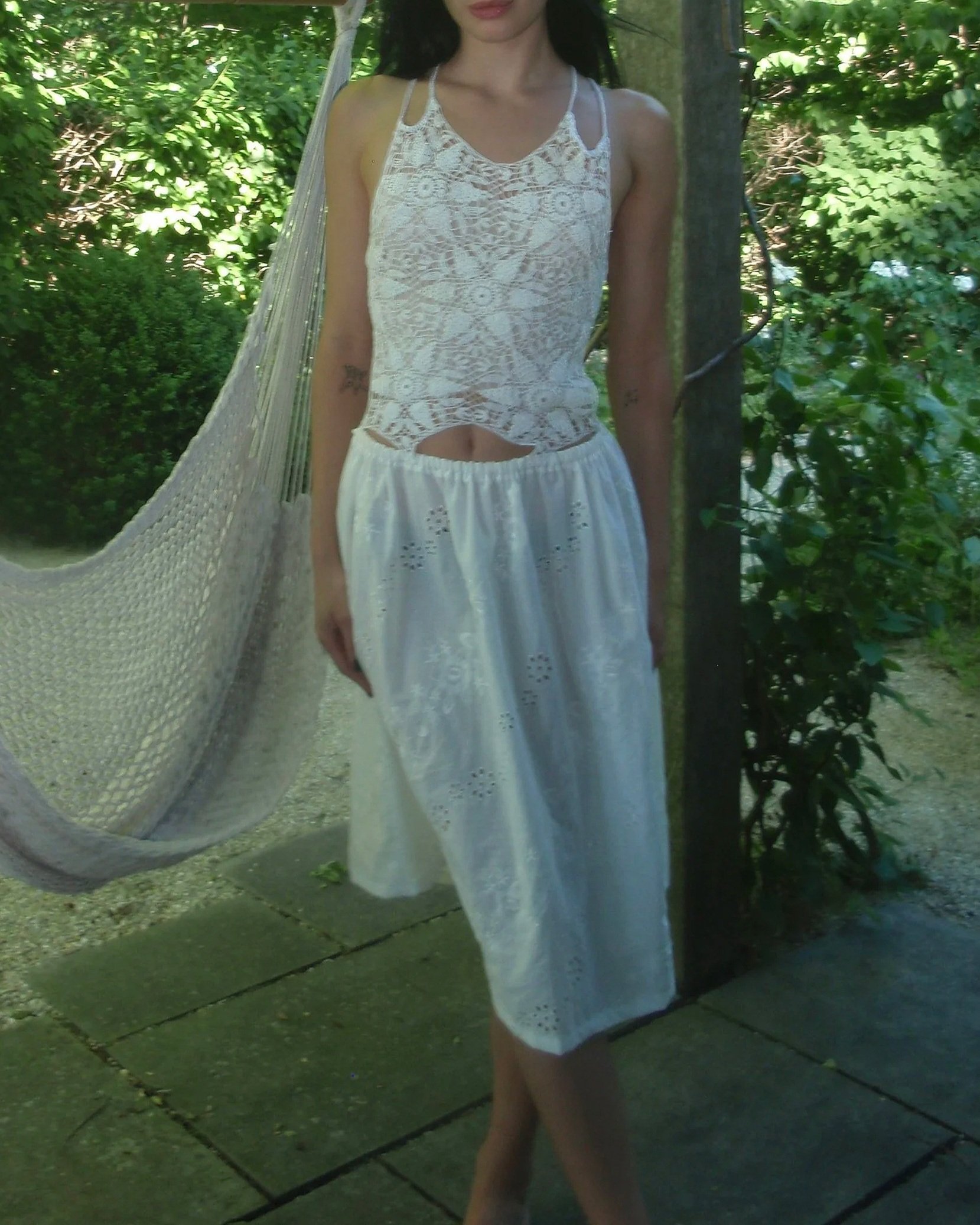 A woman wearing a white lace top and white skirt, walking outdoors surrounded by green foliage, with a hammock hanging nearby.