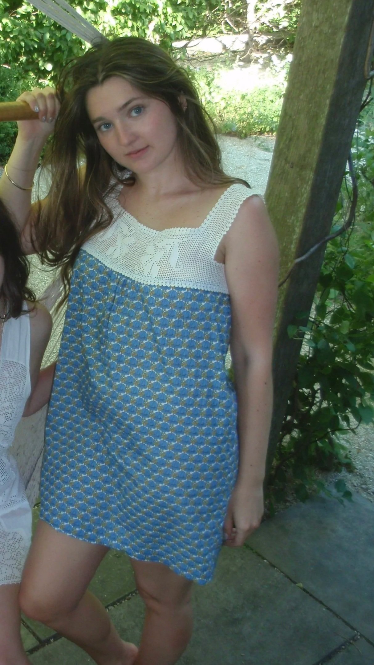 Young woman with long brown hair and blue eyes posing outdoors near a tree, wearing a sleeveless blue and white patterned dress with crochet detailing on the top, sunlight filtering through leaves.