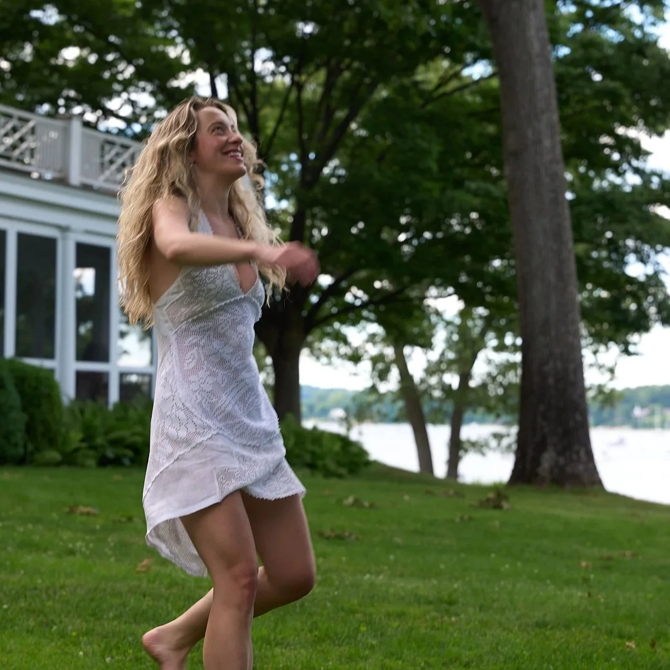 Woman in white lace dress smiling and running outside on green grass, near trees and a large house with a deck.