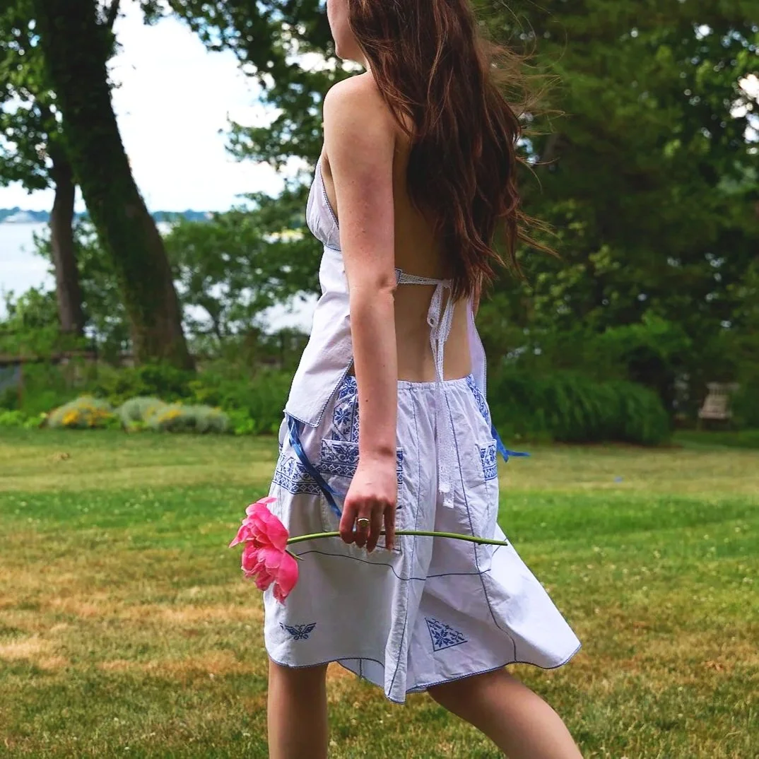 Woman with long brown hair wearing a white dress holding a pink flower in a grassy outdoor setting.