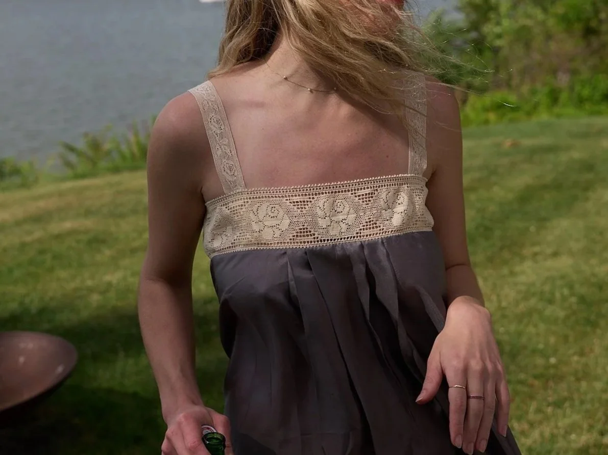 A woman in a sleeveless dress with lace detail on the top, standing outdoors near a body of water and grassy area.