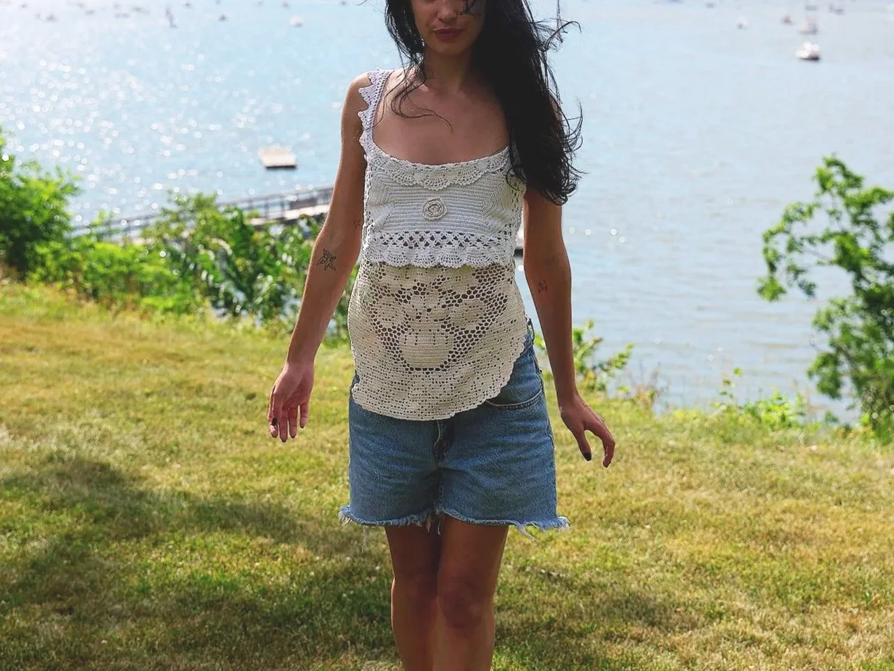 A woman in a white crocheted top and denim shorts standing outdoors near a body of water with boats, greenery, and trees in the background.