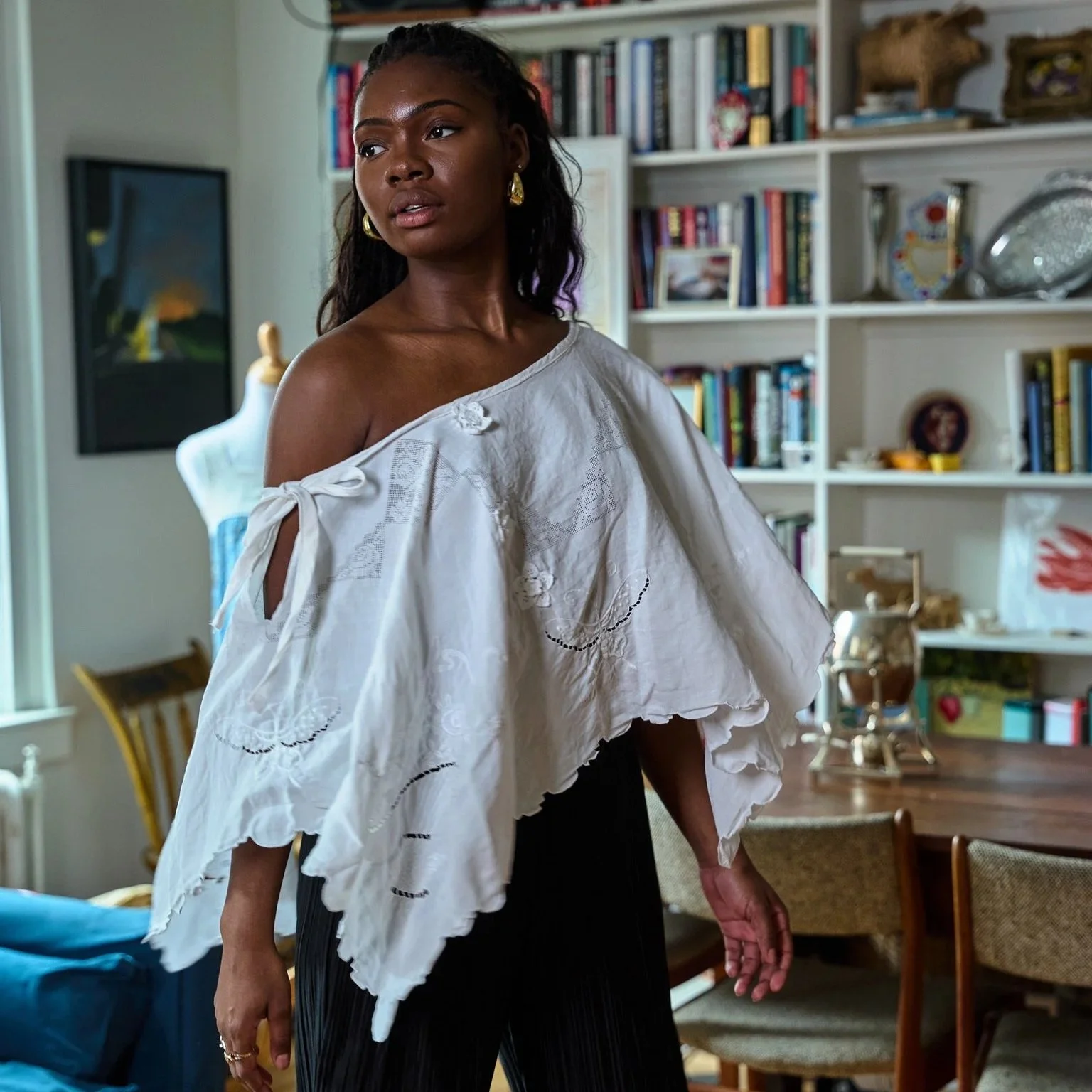 A woman with dark skin and long dark hair standing indoors in front of a bookshelf. She has gold earrings, is wearing an off-shoulder white blouse, and a black pleated skirt. The room contains various decorative items, books, and furniture.