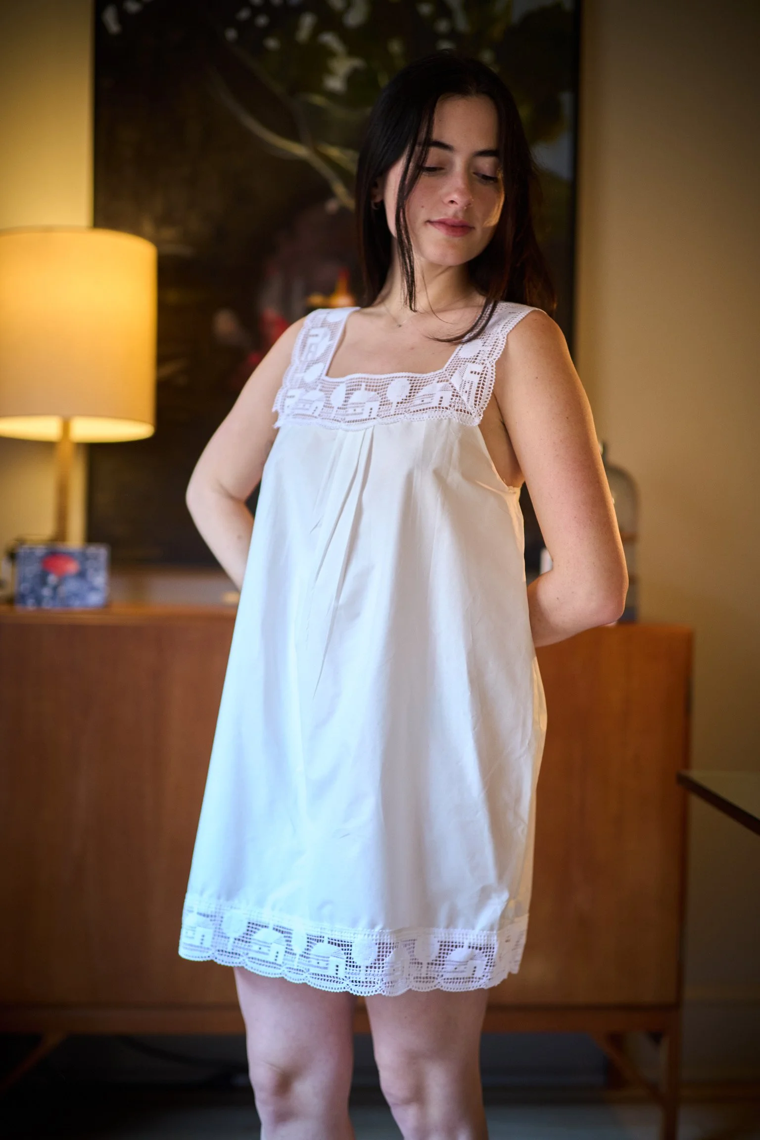 A woman with dark hair wearing a white sleeveless dress with lace details, standing in a room with a lamp and dark background. She has a serene expression, looking down with her hands behind her back.
