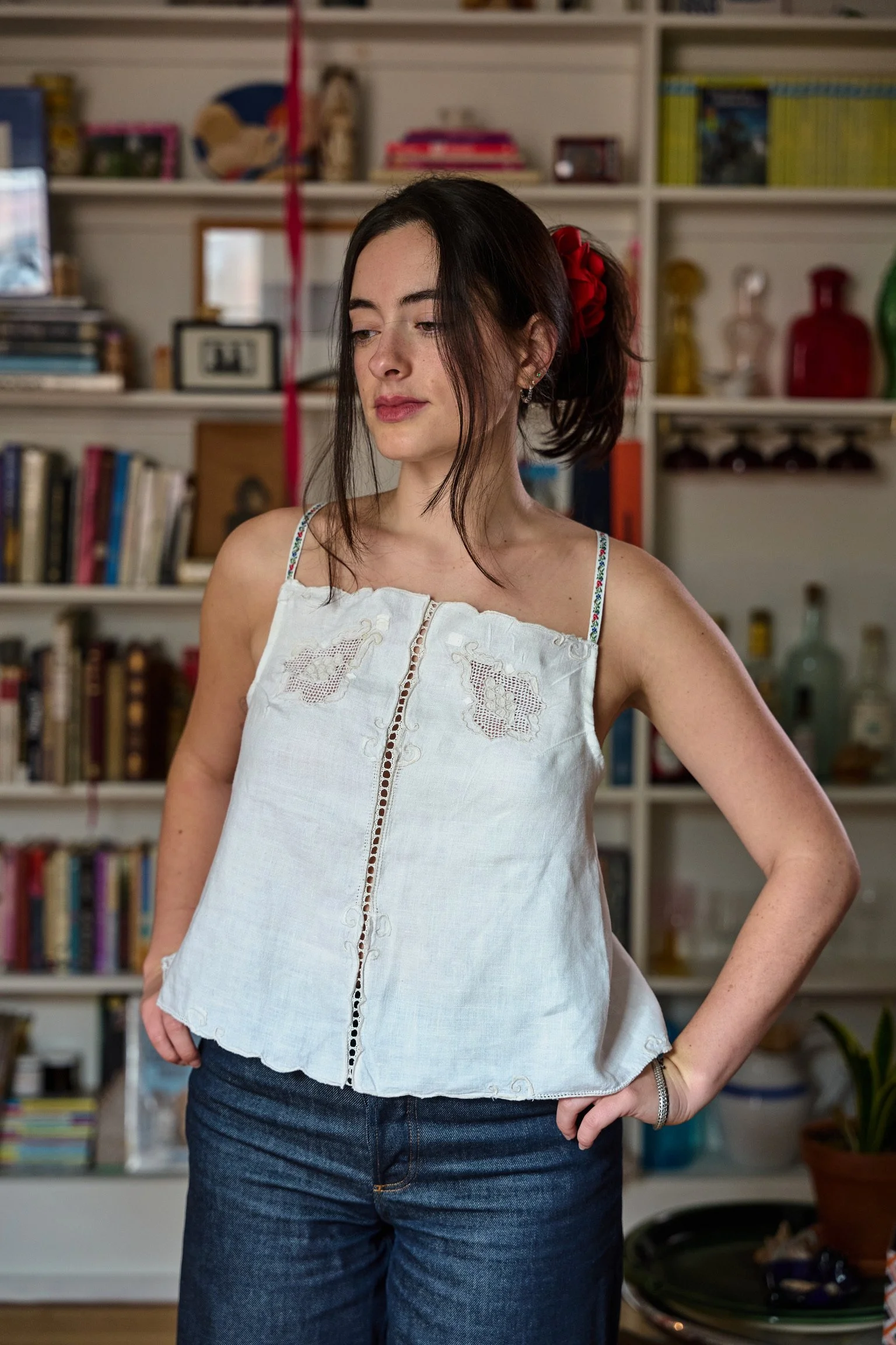 A woman with dark hair tied back with a red ribbon, wearing a white embroidered sleeveless top and dark jeans, standing in a room with a bookshelf filled with books, decorative bottles, and framed photos.