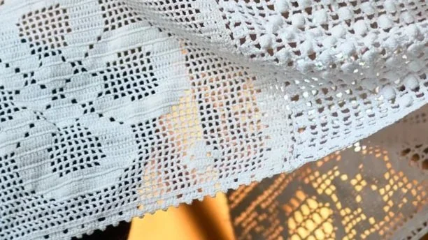 Close-up of a white lace tablecloth with intricate patterns, draped over a surface, with warm light creating shadows.