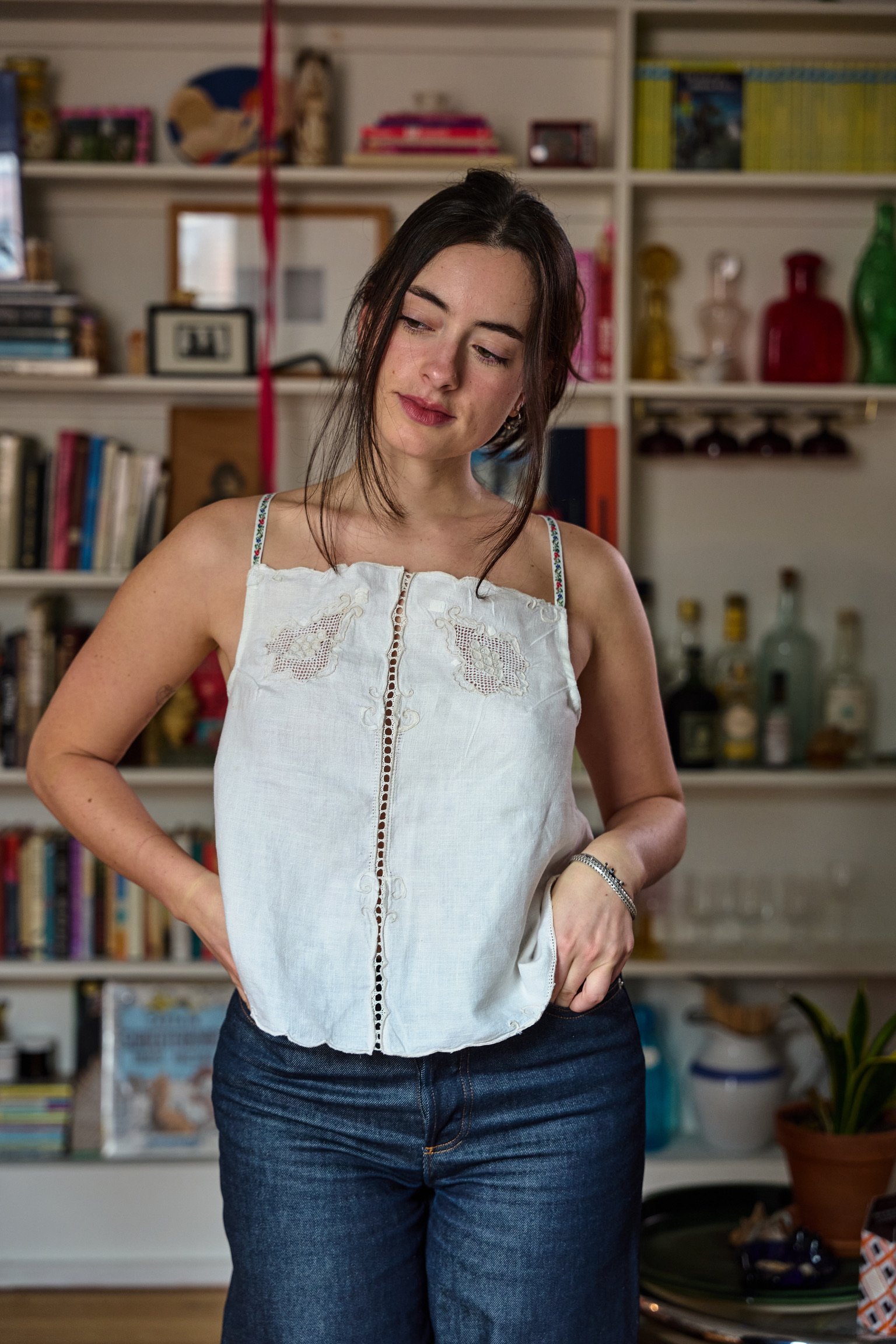 A woman with dark hair wearing a white sleeveless top and jeans, standing in a room with books and decorative items on shelves in the background.