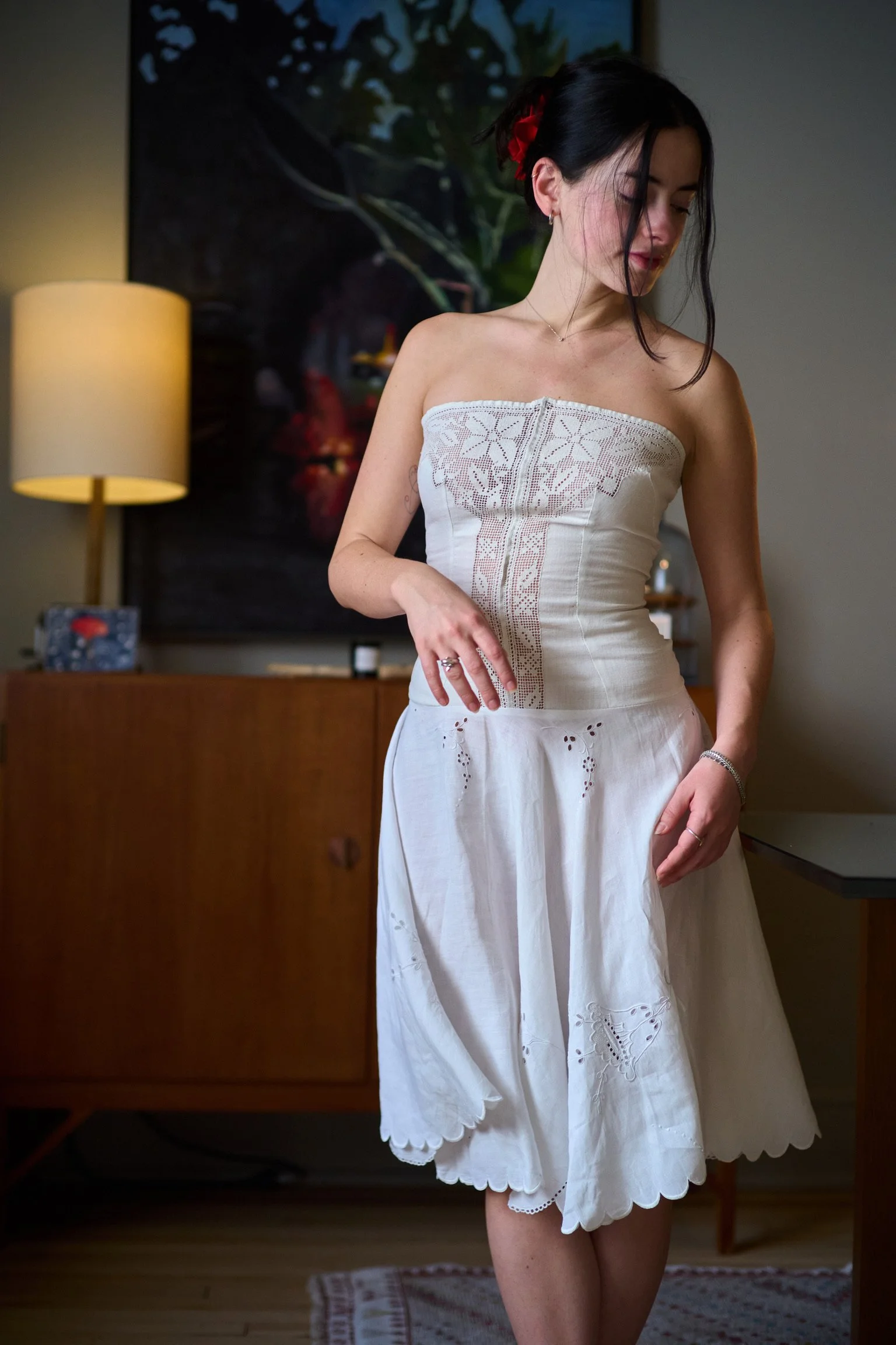 A young woman with dark hair styled with a red flower, wearing a strapless white dress with embroidery and eyelet details, standing indoors with a lamp, artwork, and wooden furniture in the background.