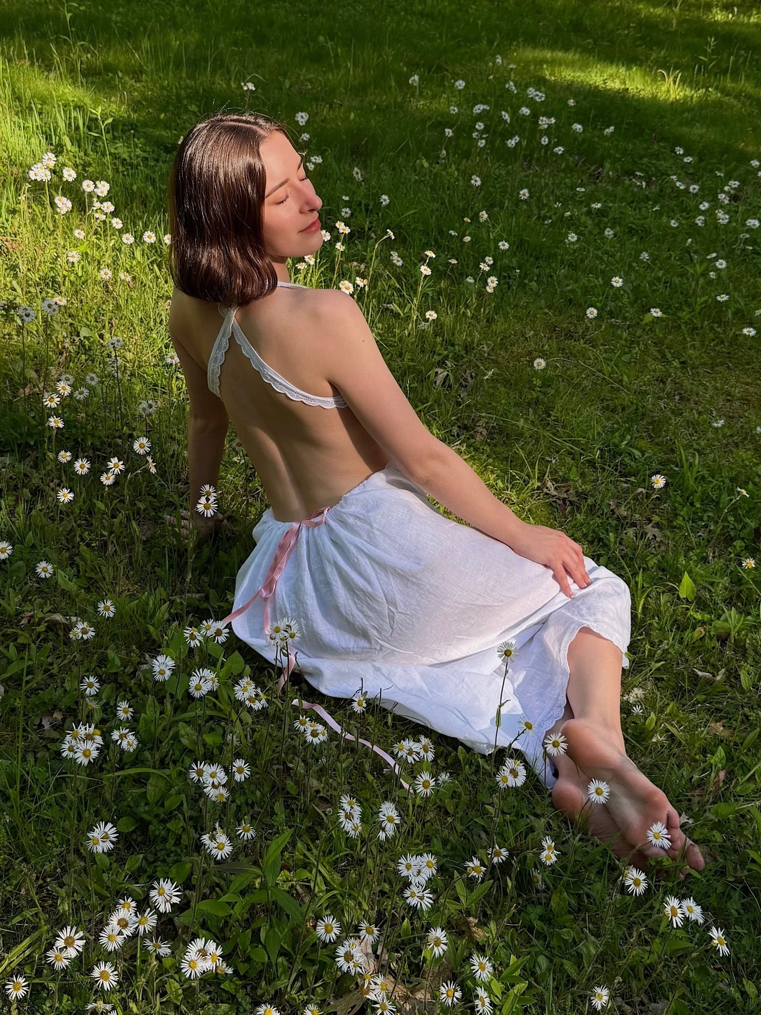 A woman with shoulder-length brown hair and light skin is sitting barefoot on a grassy field, surrounded by small white daisies. She is wearing a white dress with lace straps and has her eyes closed, appearing to enjoy the sunlight.