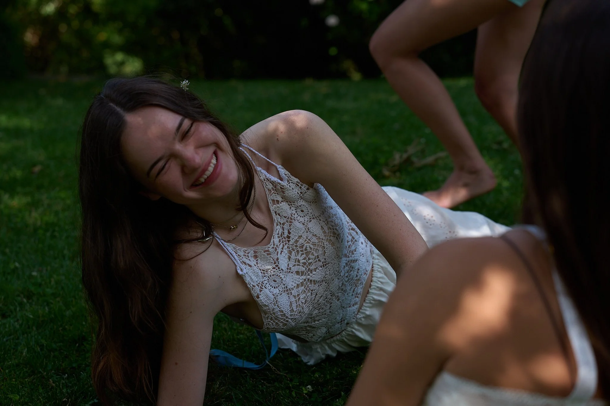 A young woman with long dark hair smiling while lying on grass, wearing a white lace sleeveless top.