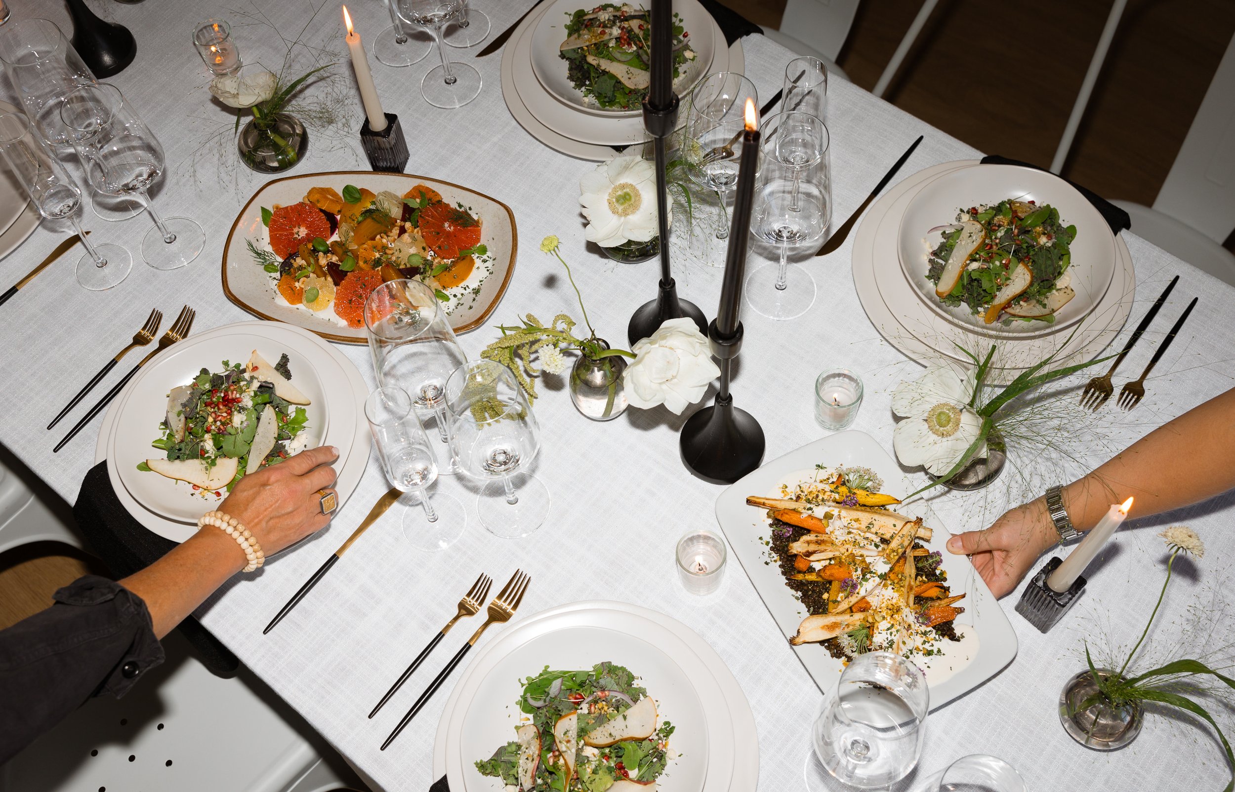 A dinner table with white tablecloth, adorned with candles and floral centerpieces, set with plates of salad and other dishes, and surrounded by empty glasses and silverware.