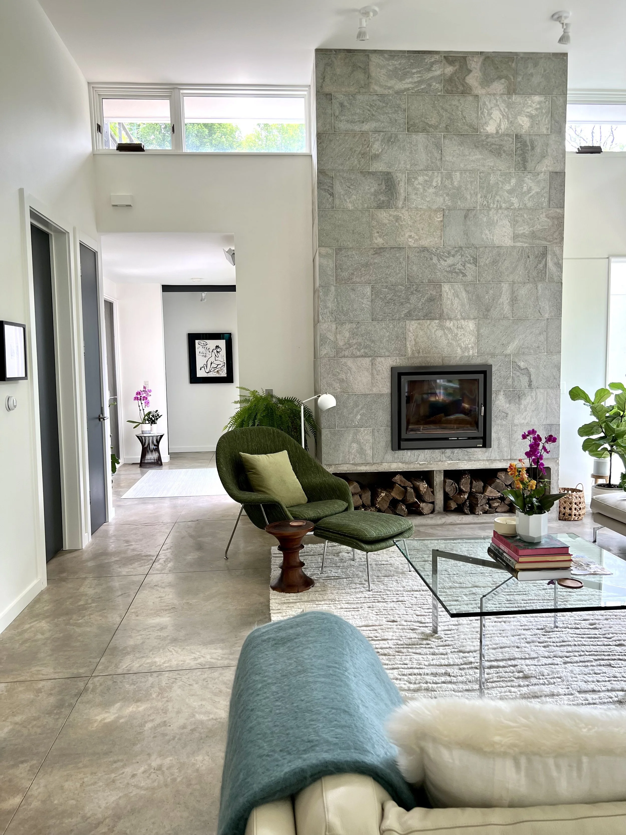 Living room with a fireplace, green chair with a cushion, glass coffee table with books and plants, and a view of a hallway with artwork, plants, and windows.