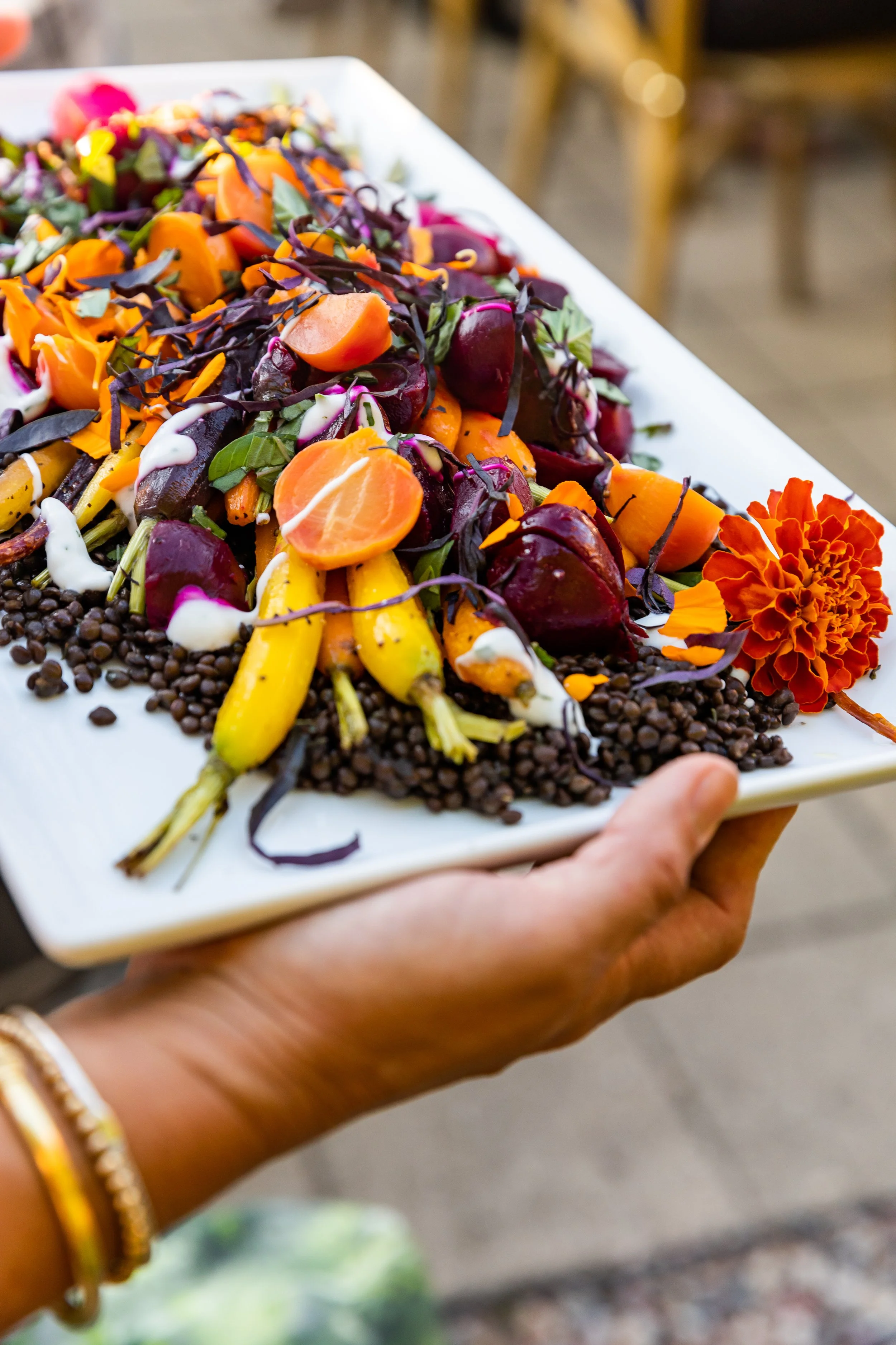 A person holding a rectangular white platter of colorful vegetables, including baby carrots, beets, yellow squash, purple cabbage, and garnished with an orange flower, on a table with a blurred background.