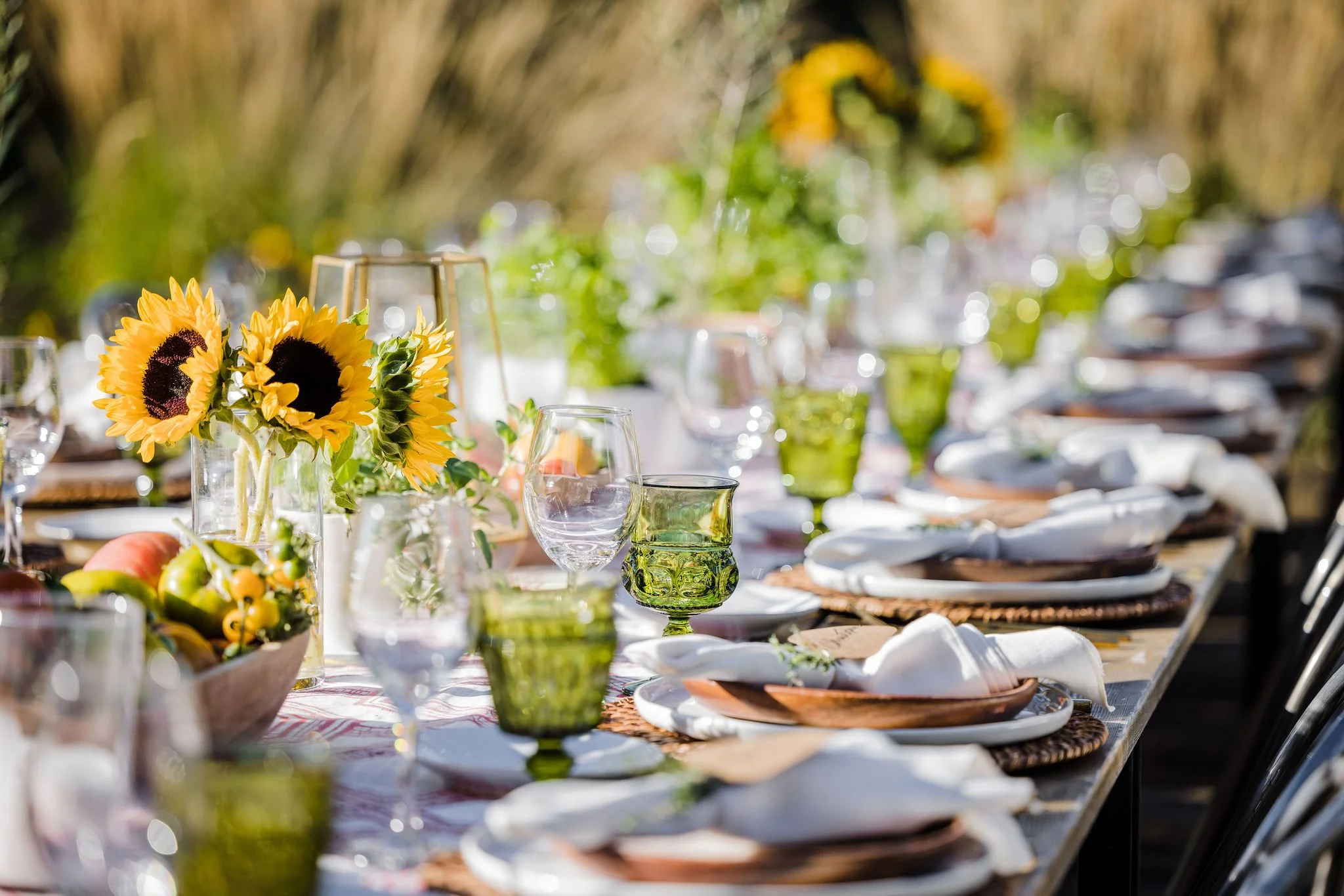 Close-up of a table set with plates, glasses, and a floral centerpiece of sunflowers, outdoors, with blurred background.