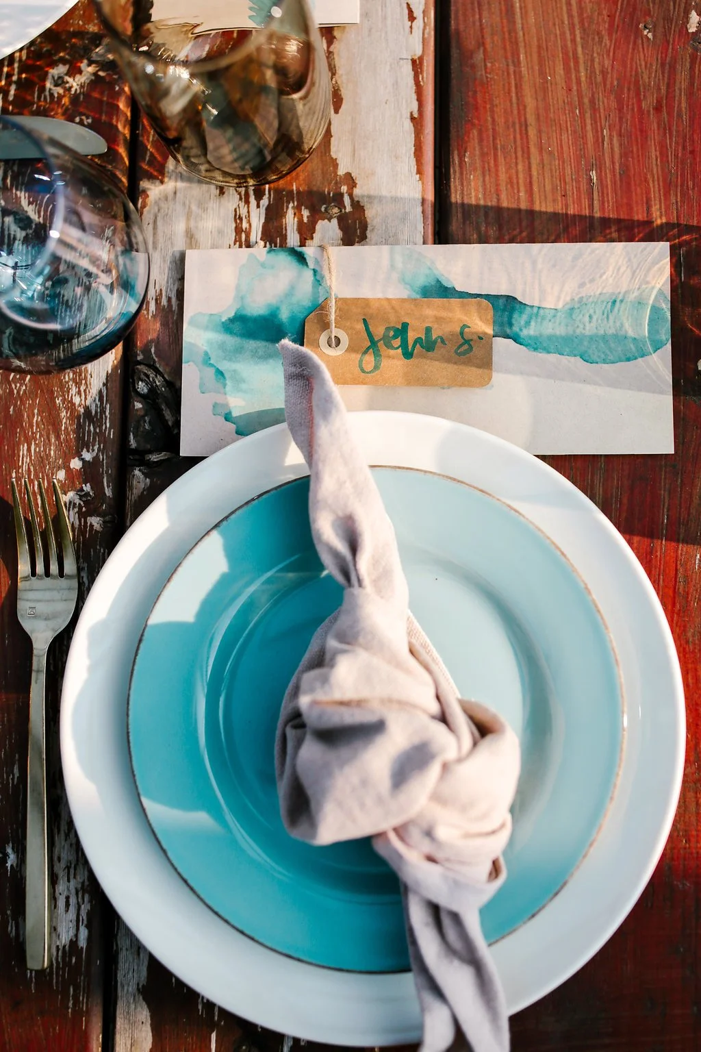 A place setting with a white plate, a turquoise bowl, a cloth napkin tied in a knot, and a fork on a rustic wooden table. There are two glasses of red wine and a watercolor place card labeled 'John S.'